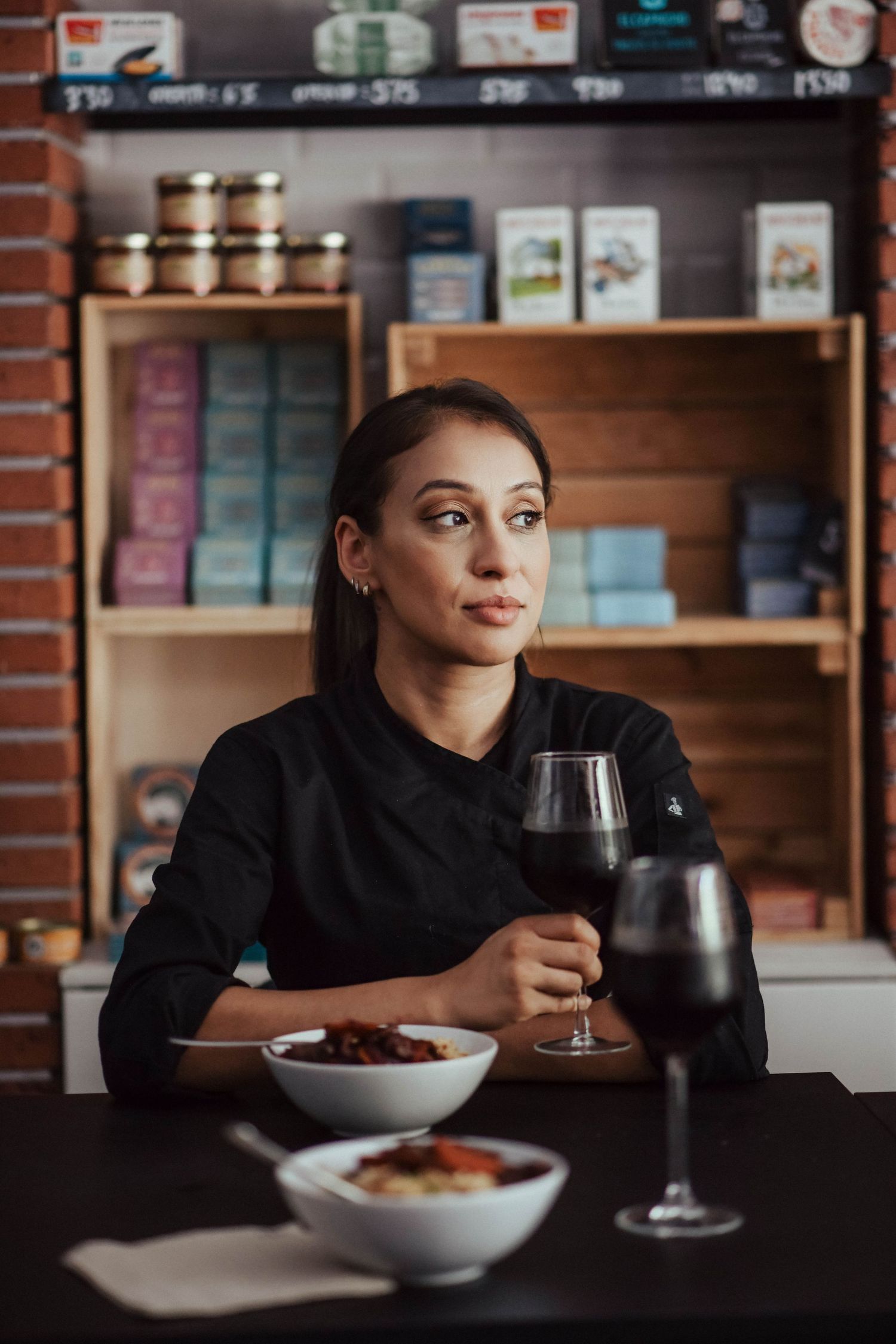 Chef in black uniform enjoying wine and food while sitting at restaurant table with shelves of cookbooks in background.