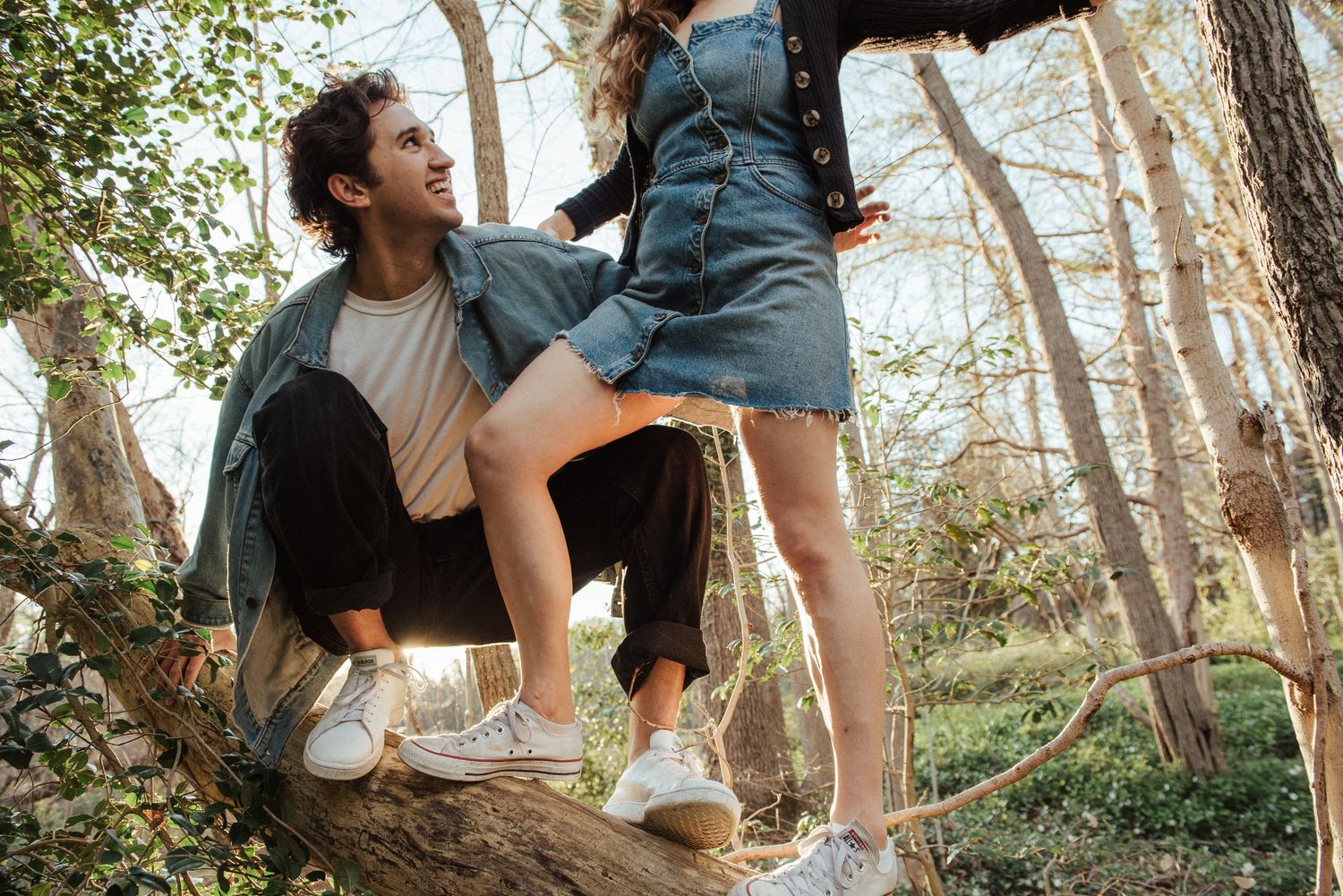 A couple sits together in a wooded area during an outdoor engagement photo session.