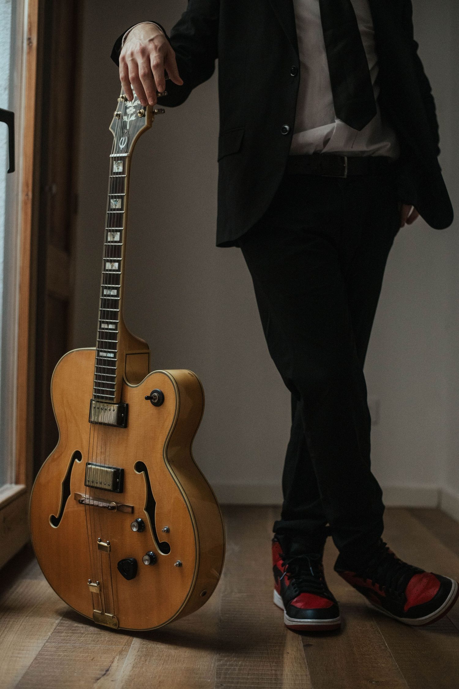 A person in black pants stands beside a vintage acoustic guitar leaning against a wall in a hallway.