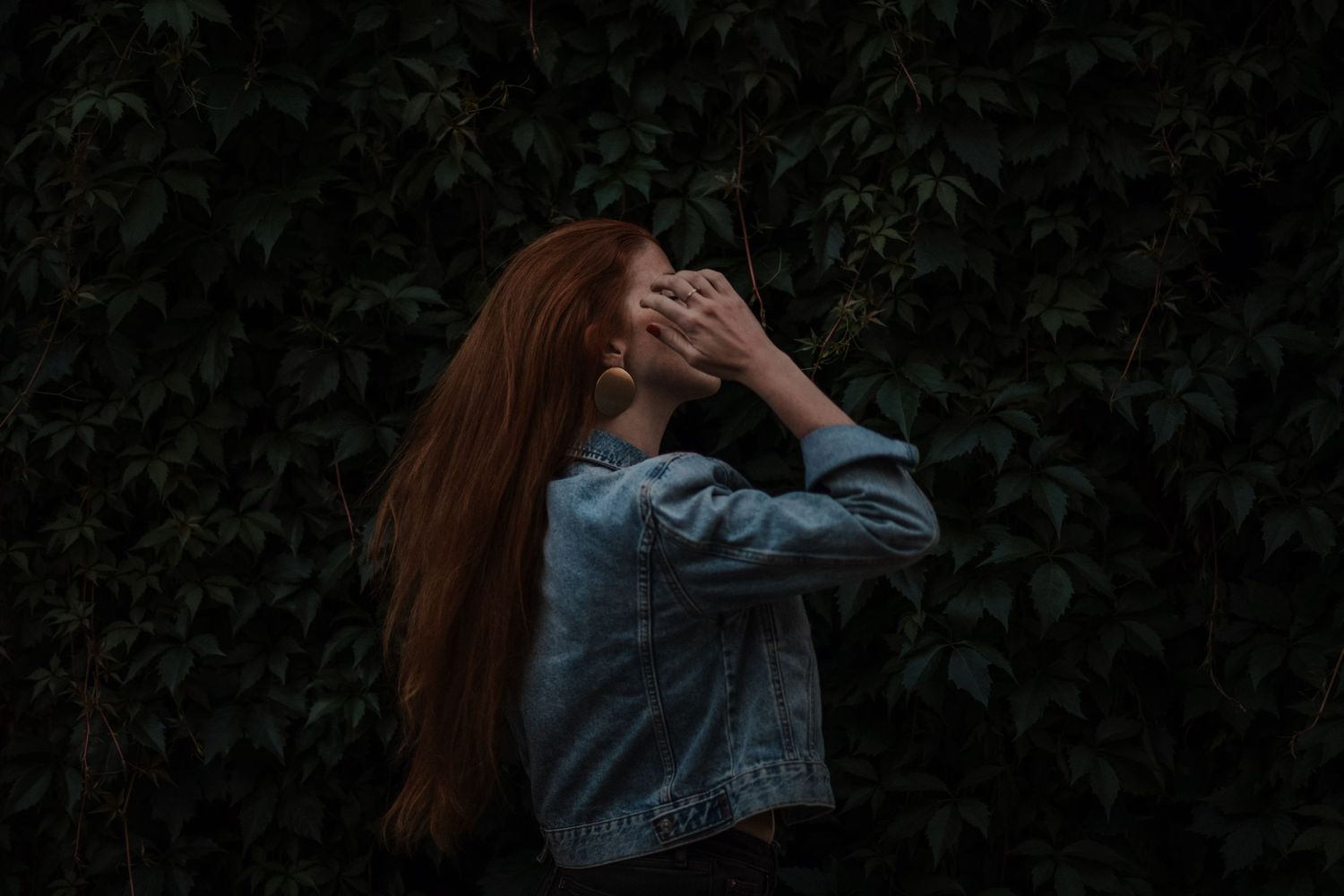 Person with long red hair wearing denim jacket stands against dark green foliage background.