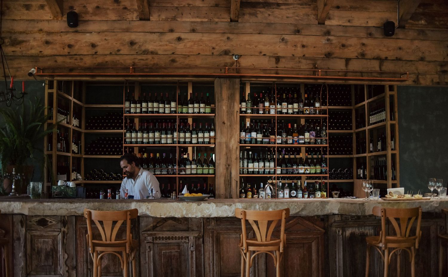 Rustic wooden bar with vintage stools and shelves displaying liquor bottles in a cozy pub setting.