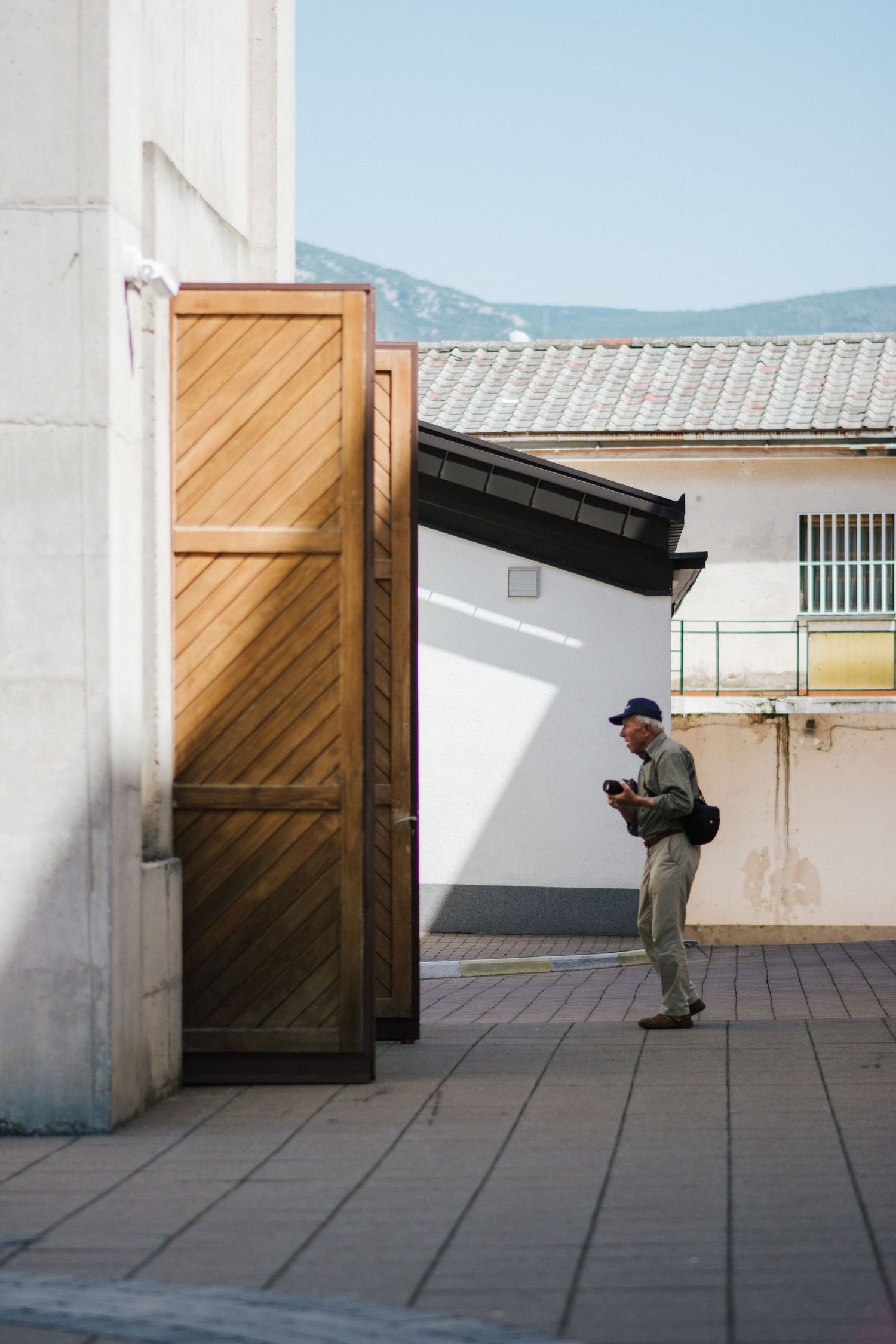 Elderly man checking camera near large wooden doors on a sunlit European street with mountains in background.