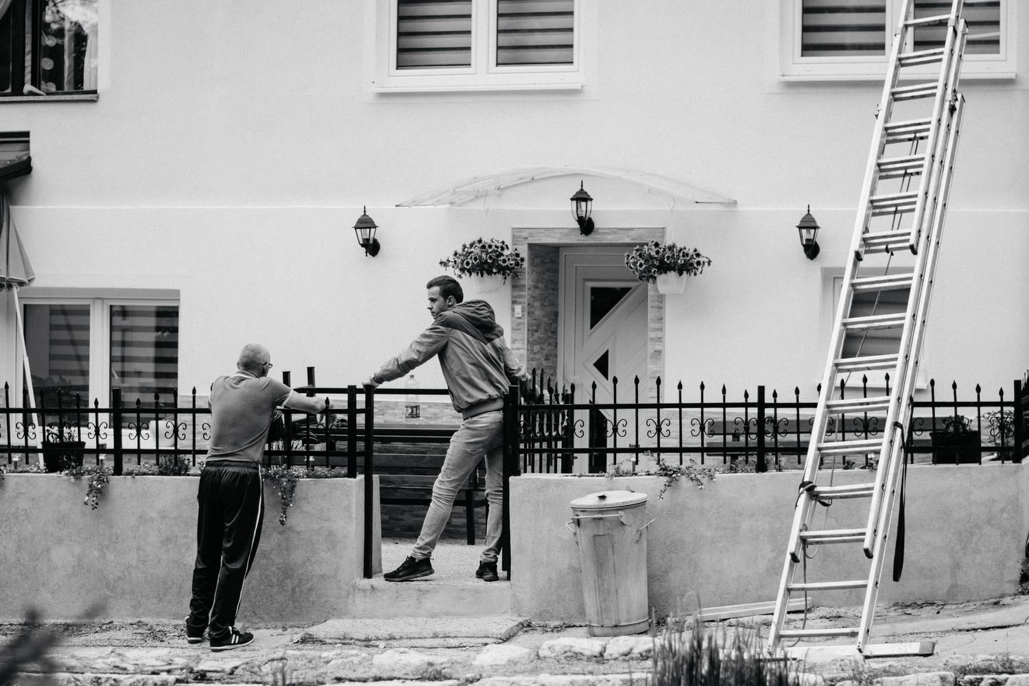 Two men at a garden gate outside a white house, with a ladder leaning against the wall in black and white.