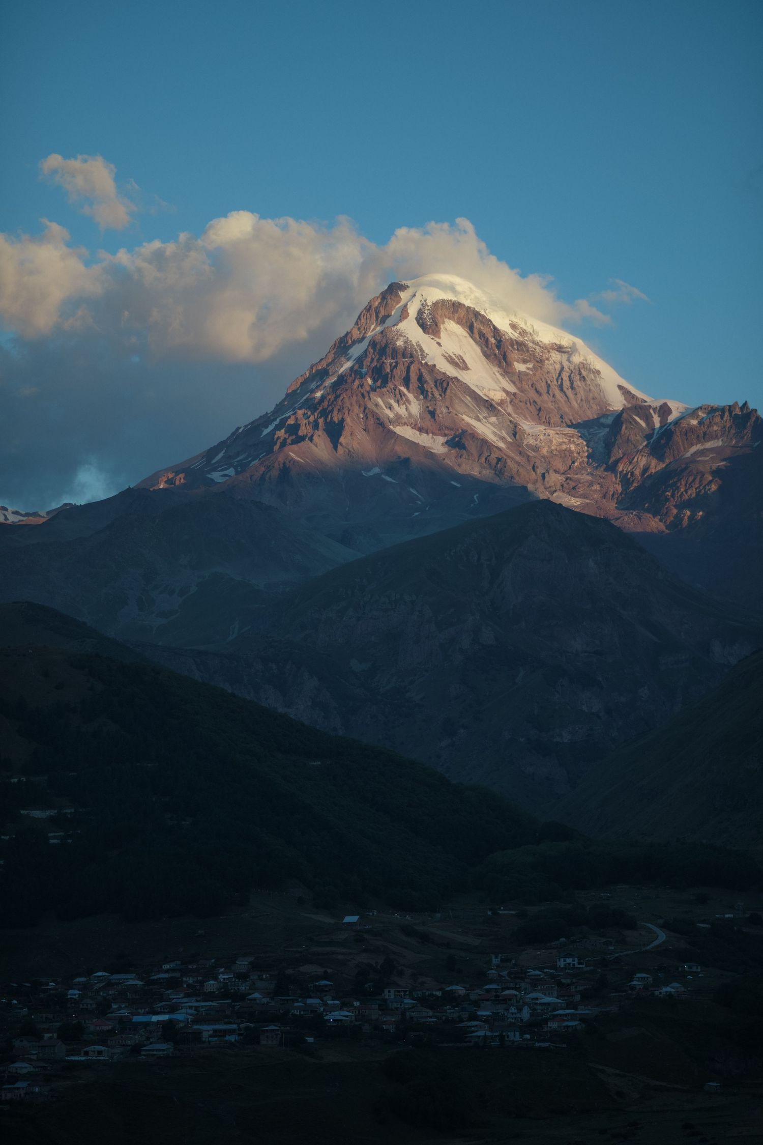 Snow-capped Mount Kazbek towers over a small village nestled in the mountains during golden hour in Georgia.