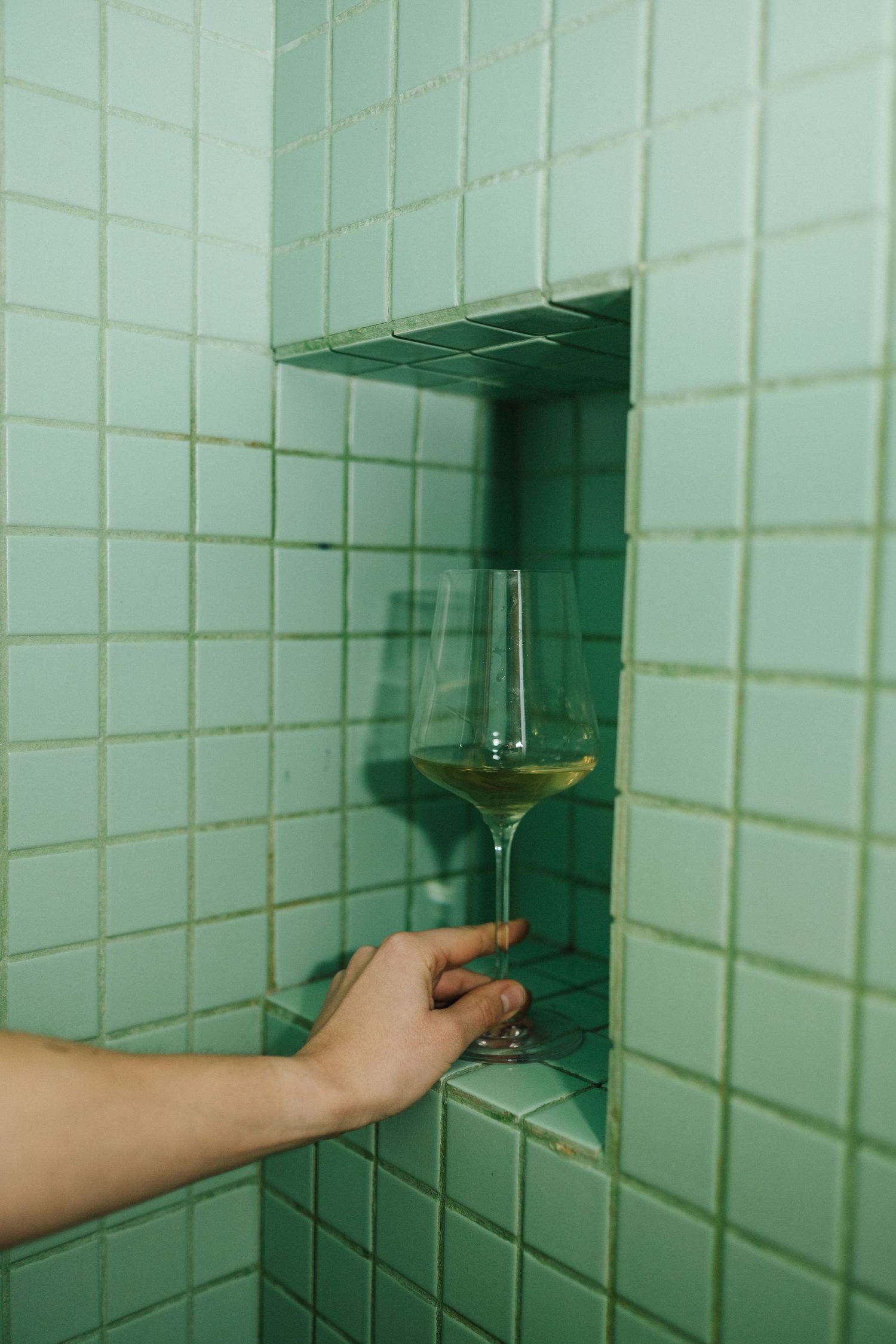 A hand placing a glass of white wine on a green tiled shower niche shelf.