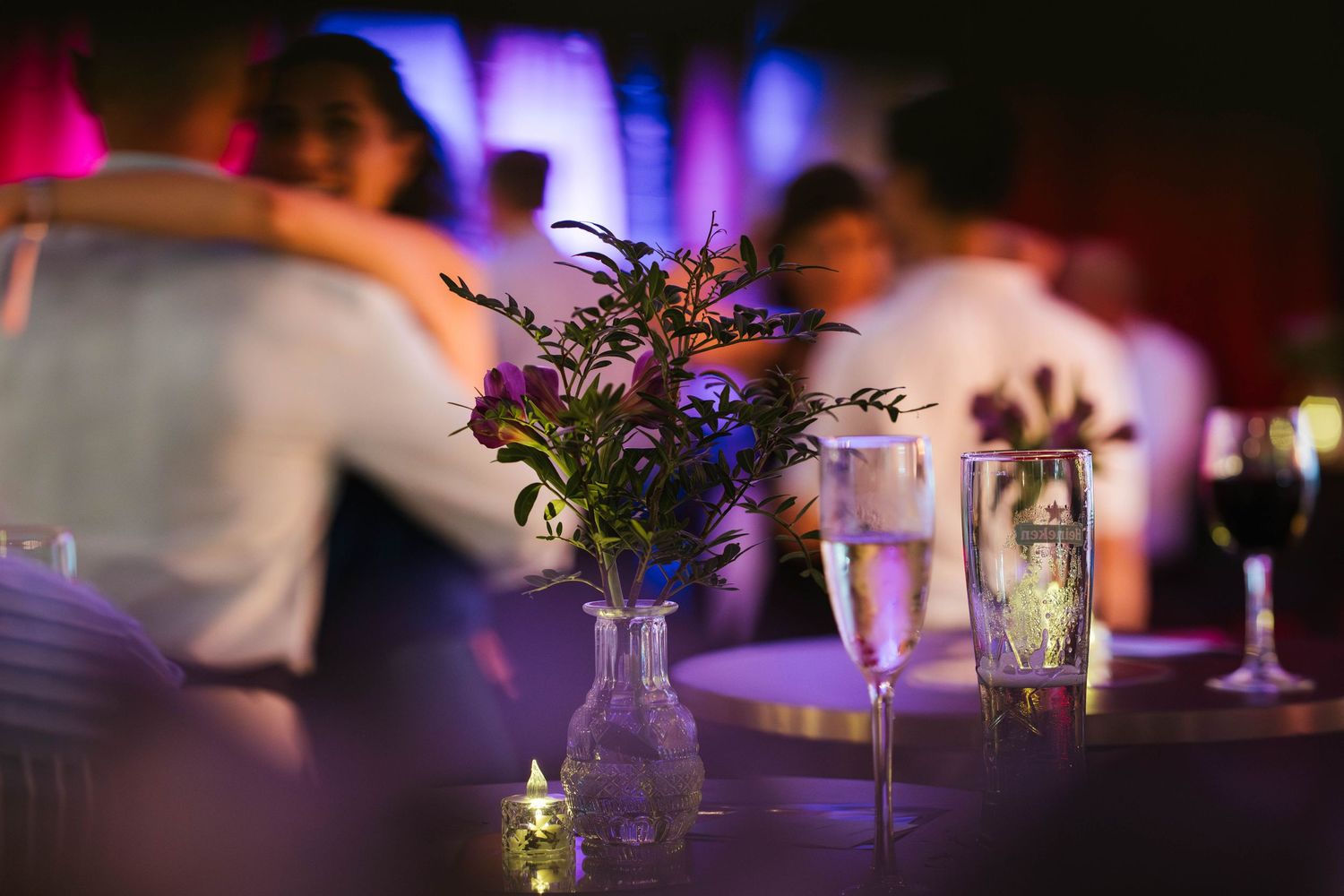 Elegant wedding reception table with purple wildflowers in glass vase, candles, and guests dancing in background.