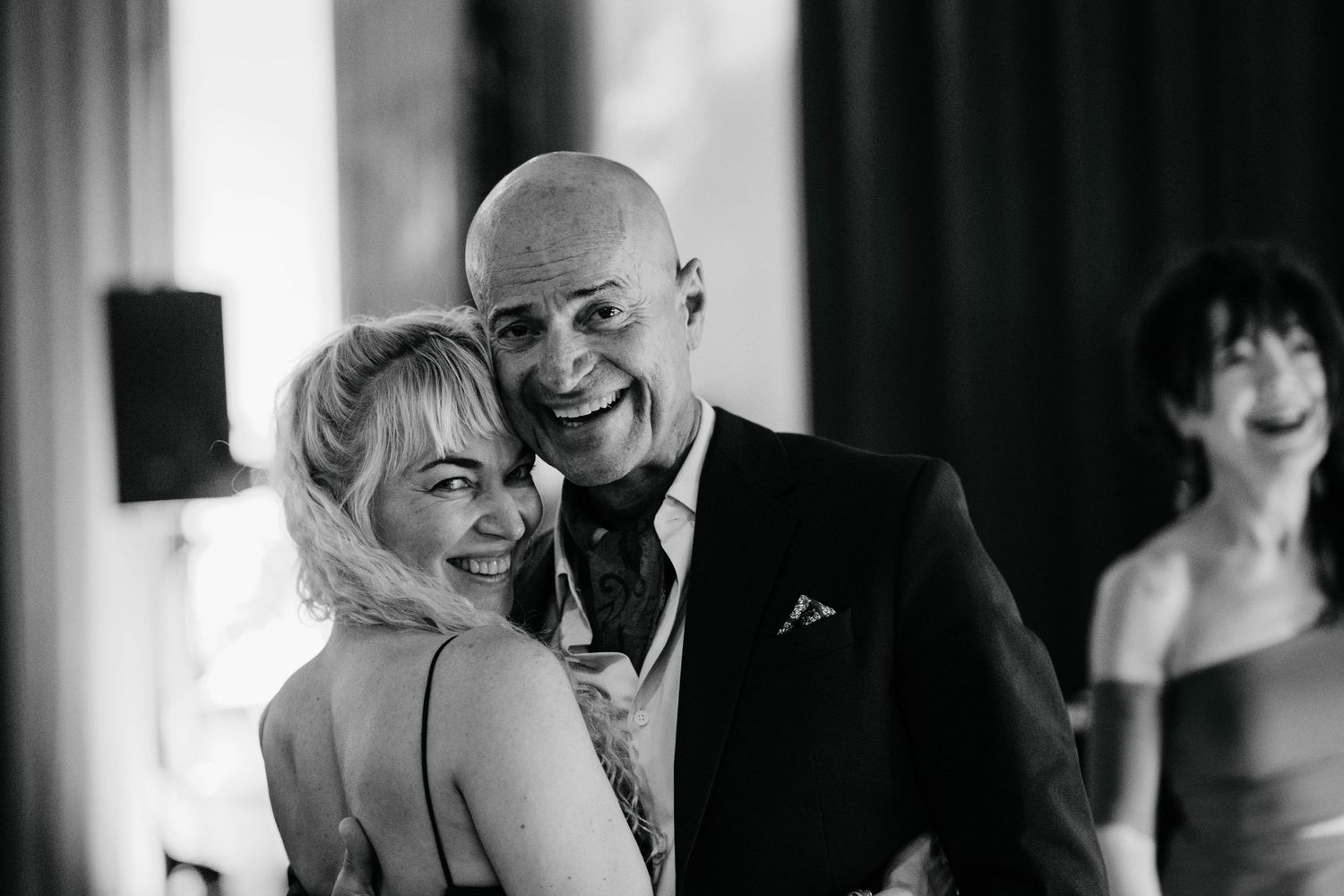 Black and white photo of a smiling bald man in a suit embracing a blonde woman at a formal event.