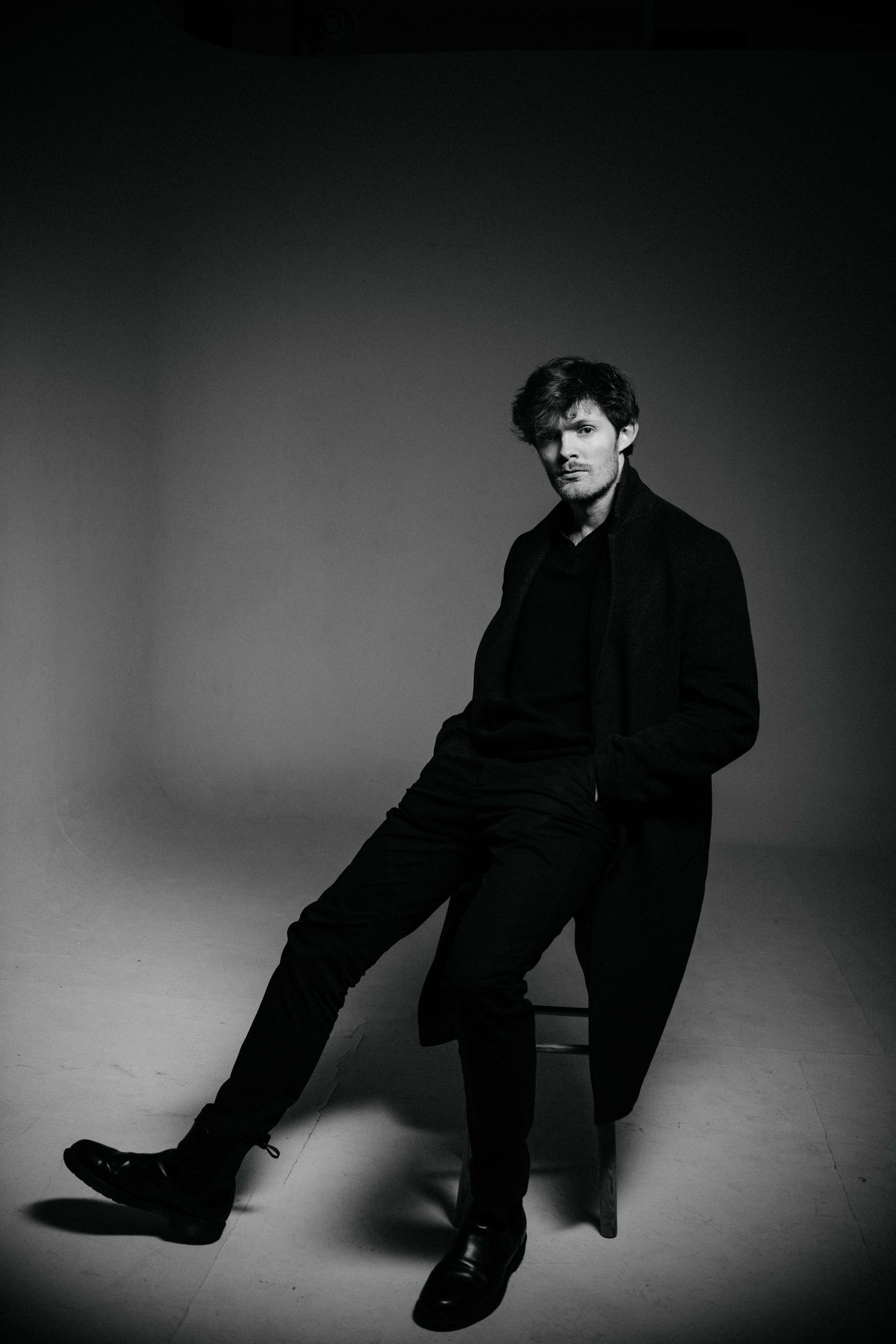 Black and white portrait of a man in a long black coat sitting on a stool in a studio setting.
