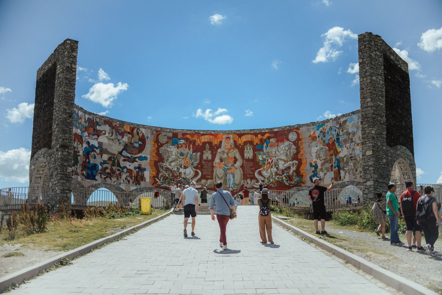 A panoramic view of tourists exploring ancient stone ruins under a bright blue sky with scattered white clouds.