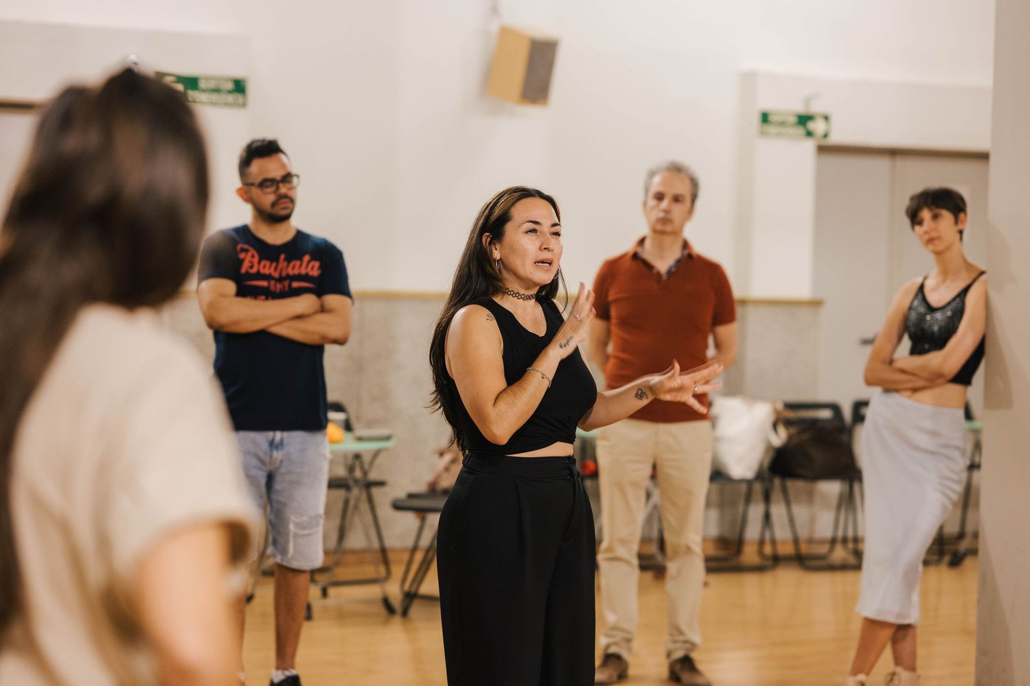 Group of people in a dance or acting rehearsal studio, with a woman in black leading the session.