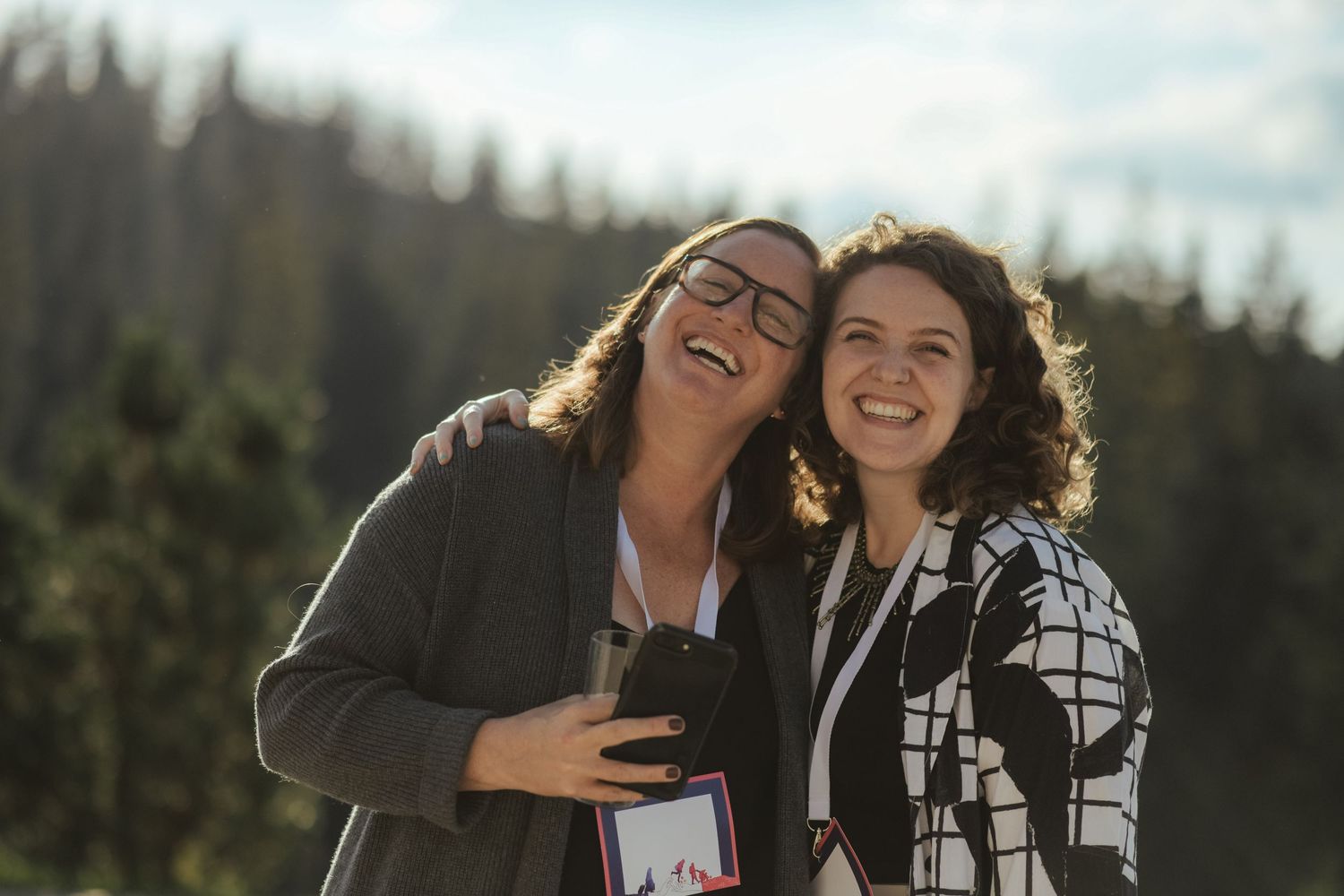 Friends laughing together outdoors with evergreen trees in the background.
