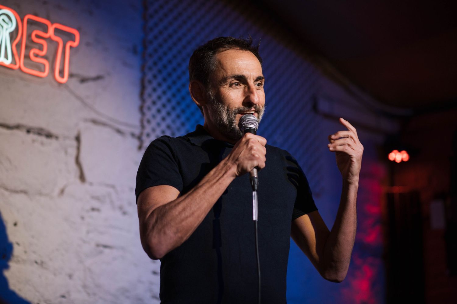 Stand-up comedian performing on stage with brick wall backdrop and moody lighting.