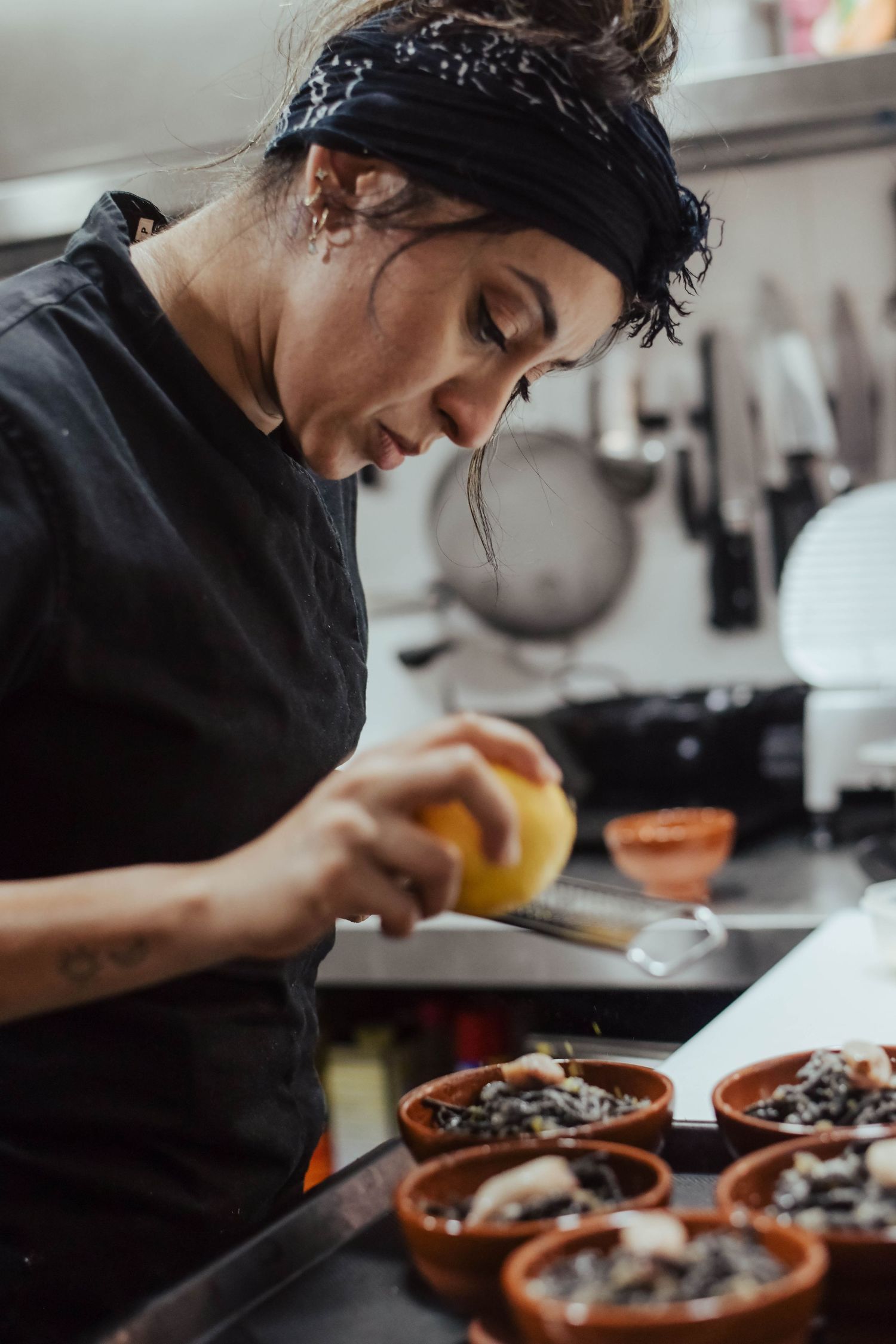 Chef in black uniform and bandana prepares dishes in terra cotta bowls in restaurant kitchen.