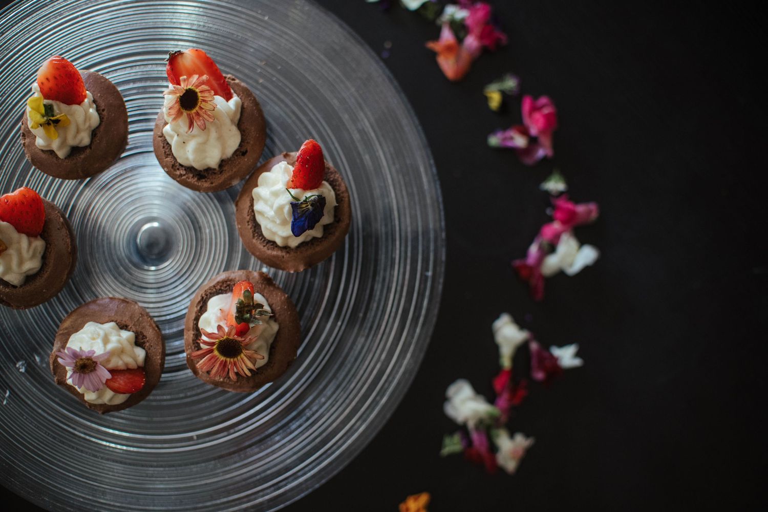 Chocolate cupcakes topped with whipped cream and fresh strawberries arranged on a dark glass plate with scattered rose petals.