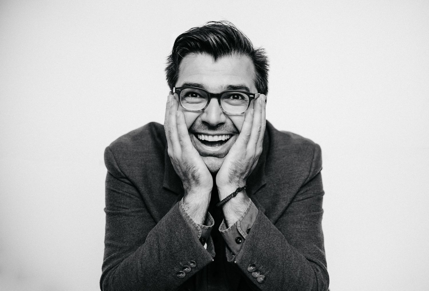 Black and white portrait of a joyful person wearing glasses with hands on cheeks and a wide smile.