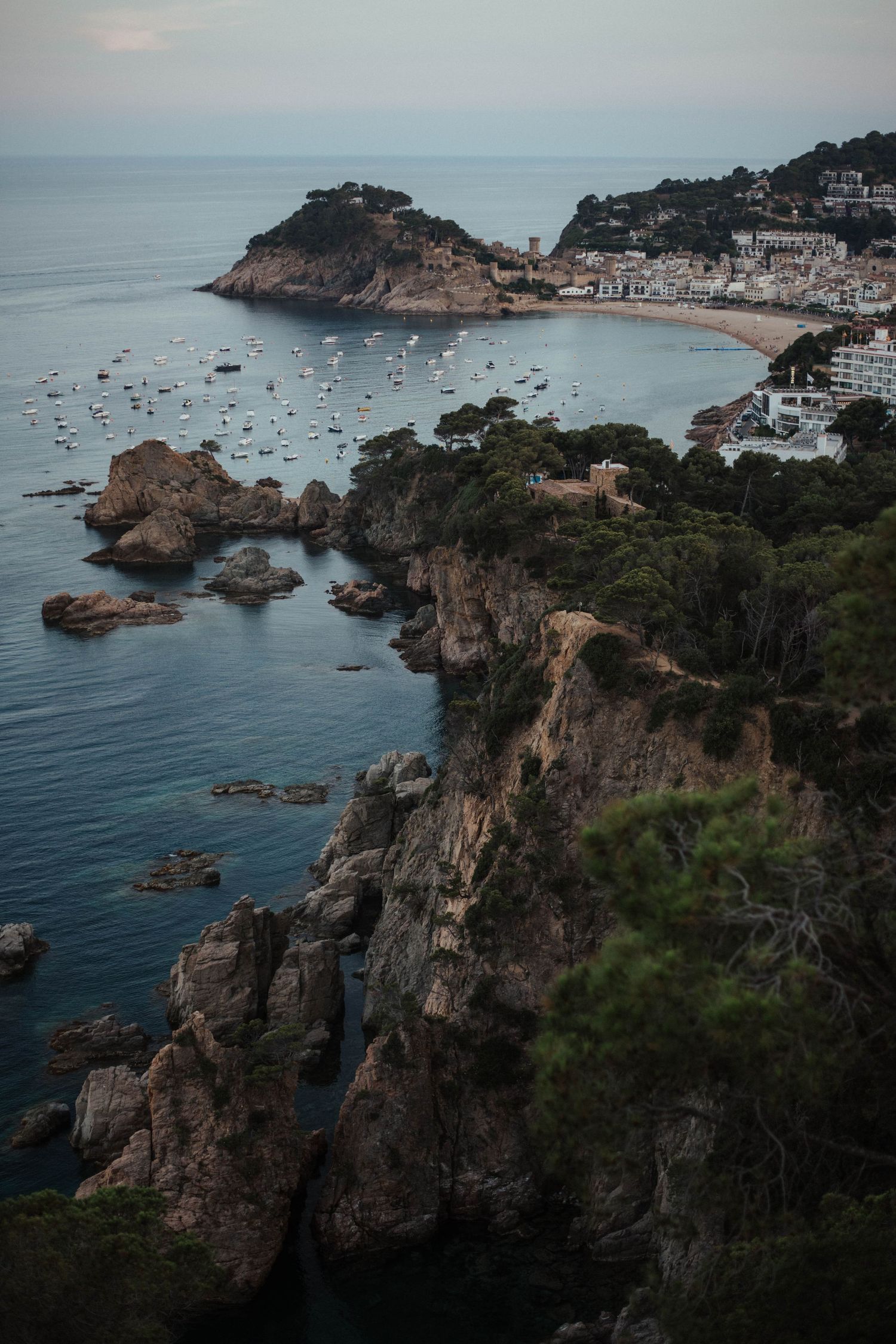 Dramatic rocky coastal cliffs overlooking the Mediterranean Sea with small town and boats visible in the distance at sunset.