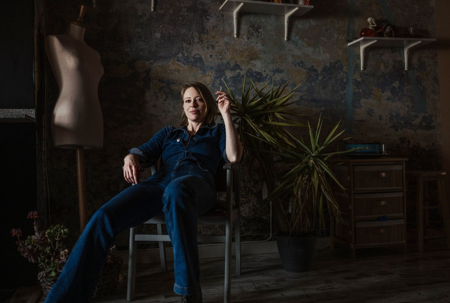 Artist relaxing in a chair in a moody studio with a dress form, plant, and textured blue walls.