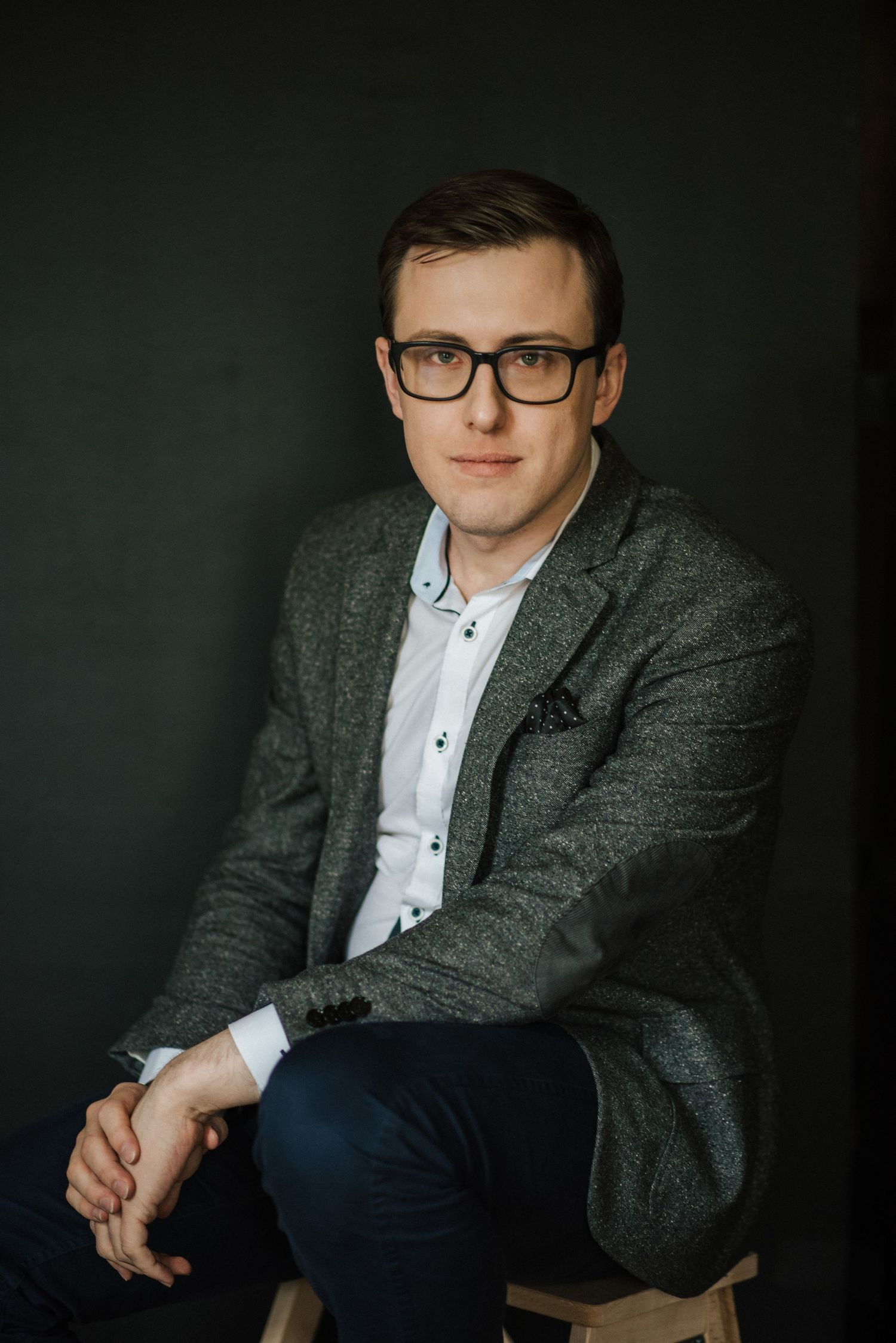 Professional studio portrait of person in glasses wearing gray tweed blazer and white shirt against dark background.