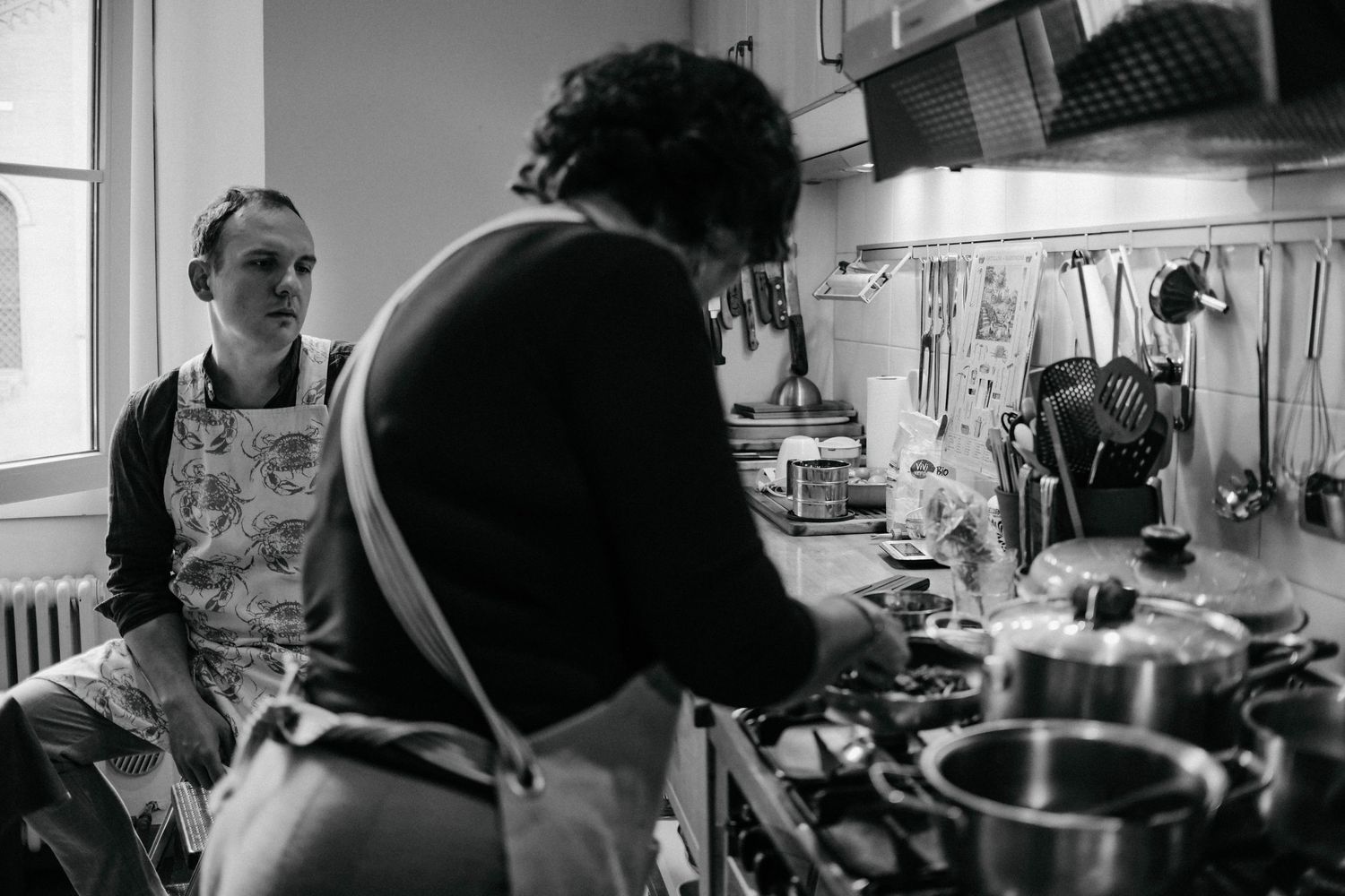 A black and white photo shows two people working at making pasta at wooden tables in an Italian kitchen.