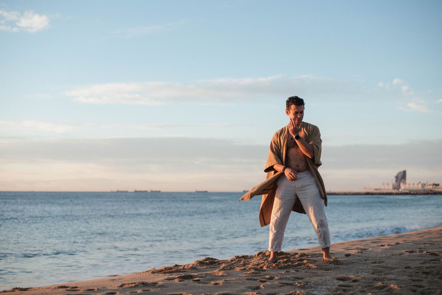 Someone poses on a sandy beach at sunset with the ocean and distant buildings visible in the background.