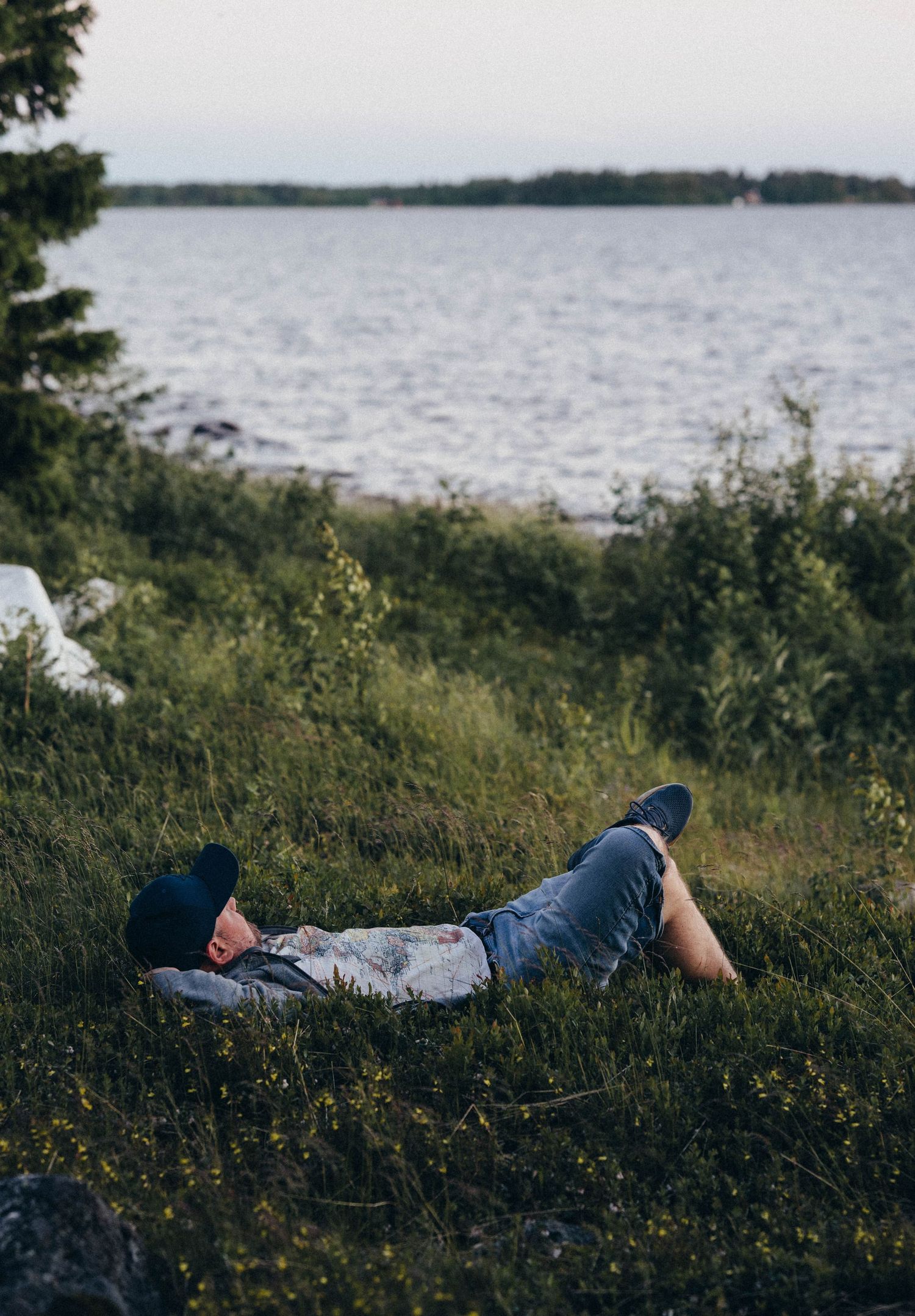 A person wearing a cowboy hat relaxes lying on green grass by a scenic lakeside shore.