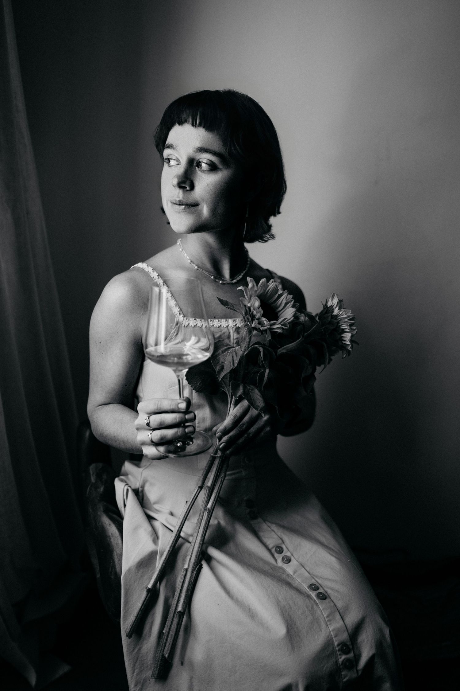A black and white portrait of someone in a light dress holding flowers and a glass, with dramatic lighting.