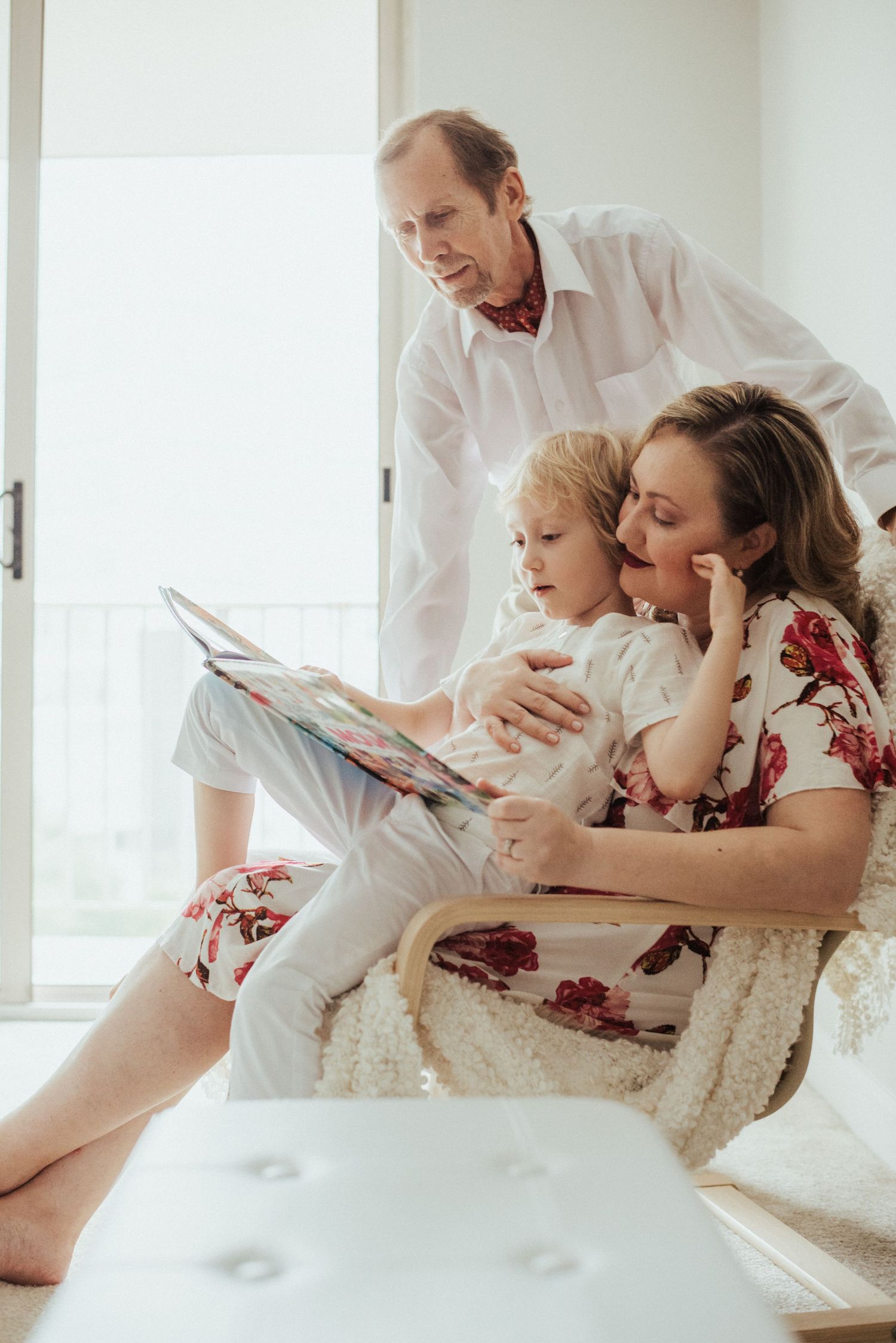 Family sharing a tender moment while reading together on a white couch in a sunlit room.
