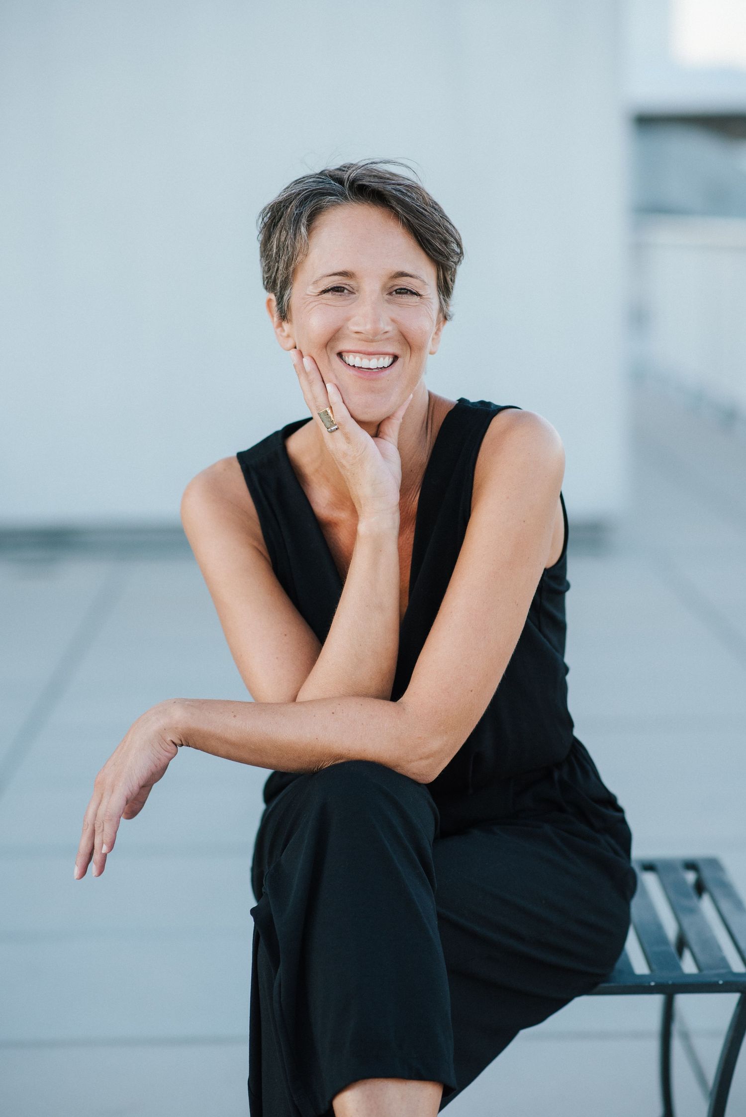 Professional portrait of someone in a black sleeveless outfit sitting casually and smiling against a light background.