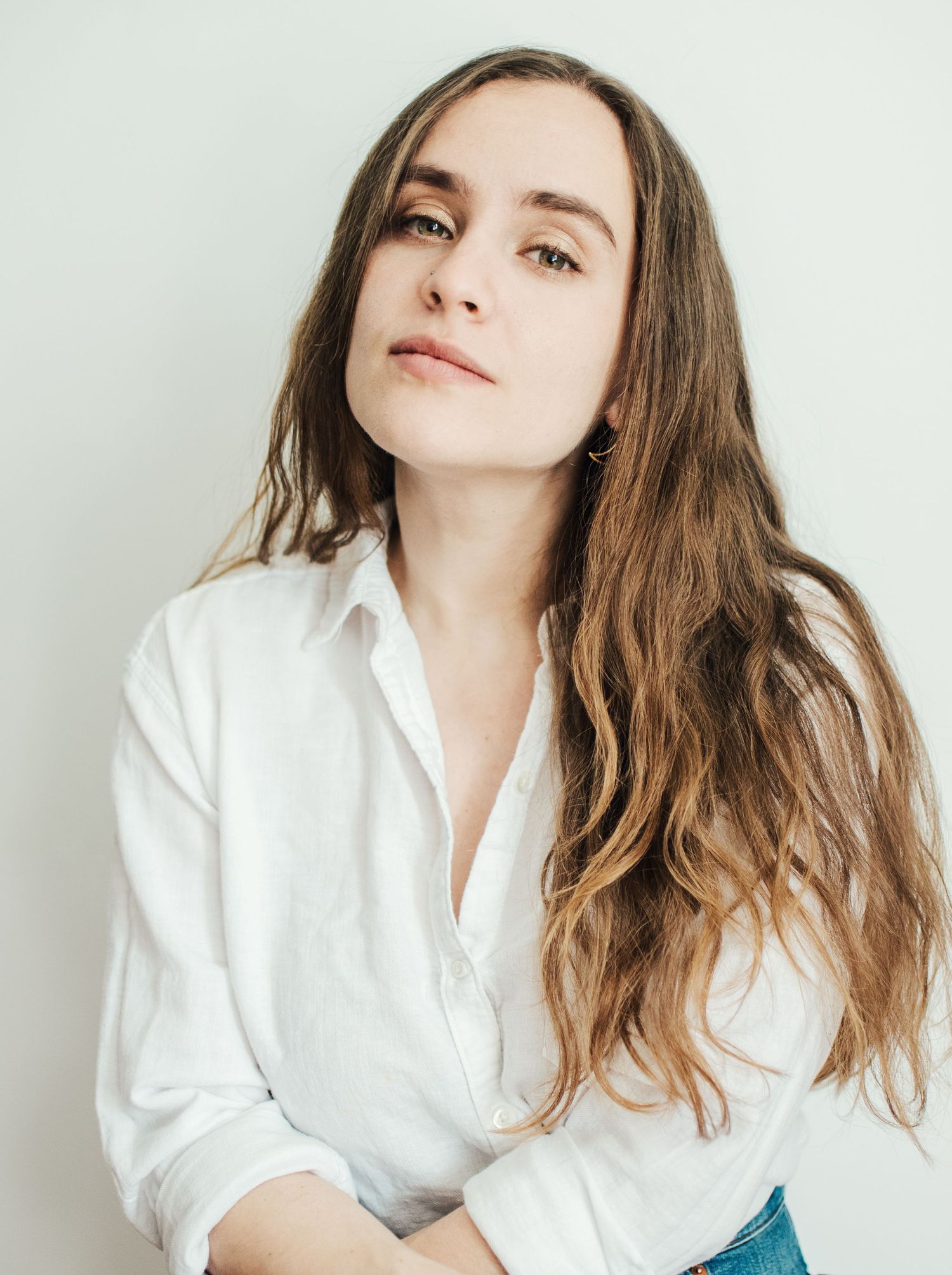 Portrait of a person with long wavy brown hair wearing a white button-down shirt against a light background.