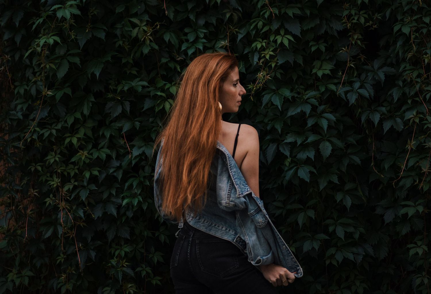 Person in denim jacket walks past green foliage wall outdoors.
