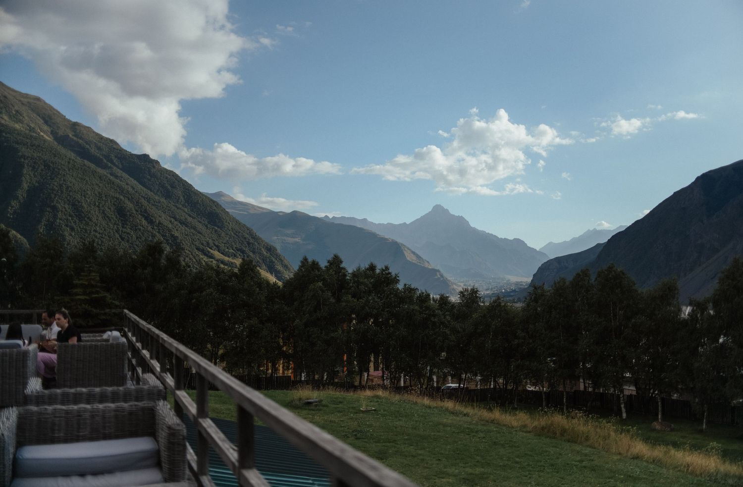 Panoramic view of snow-capped mountain peaks under cloudy skies with green valley and forest in the foreground.