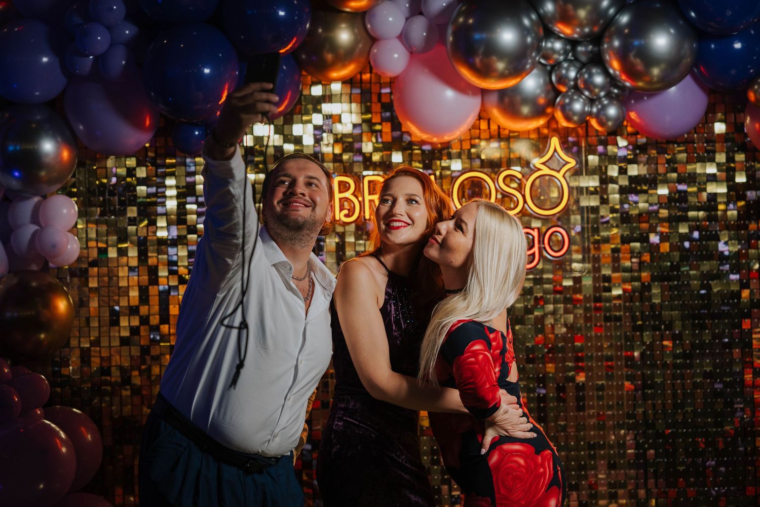 Three friends posing and smiling at a festive party with gold sequin backdrop and colorful balloons.