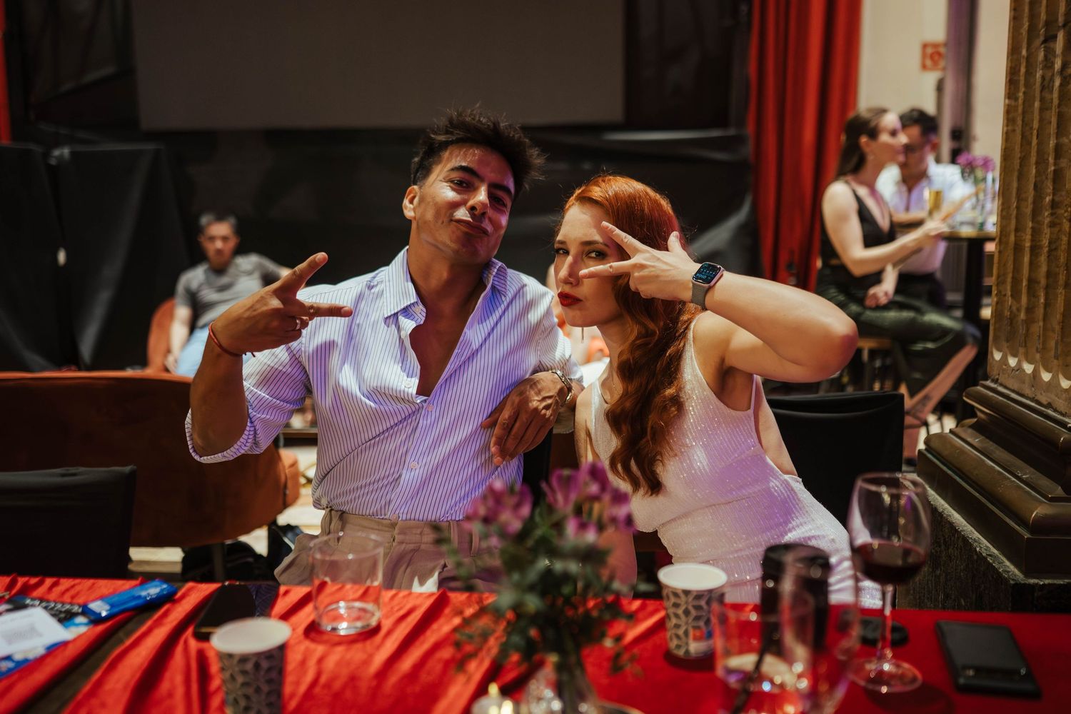Two friends pose playfully at a festive table with red decor, making peace signs and smiling at a social event.