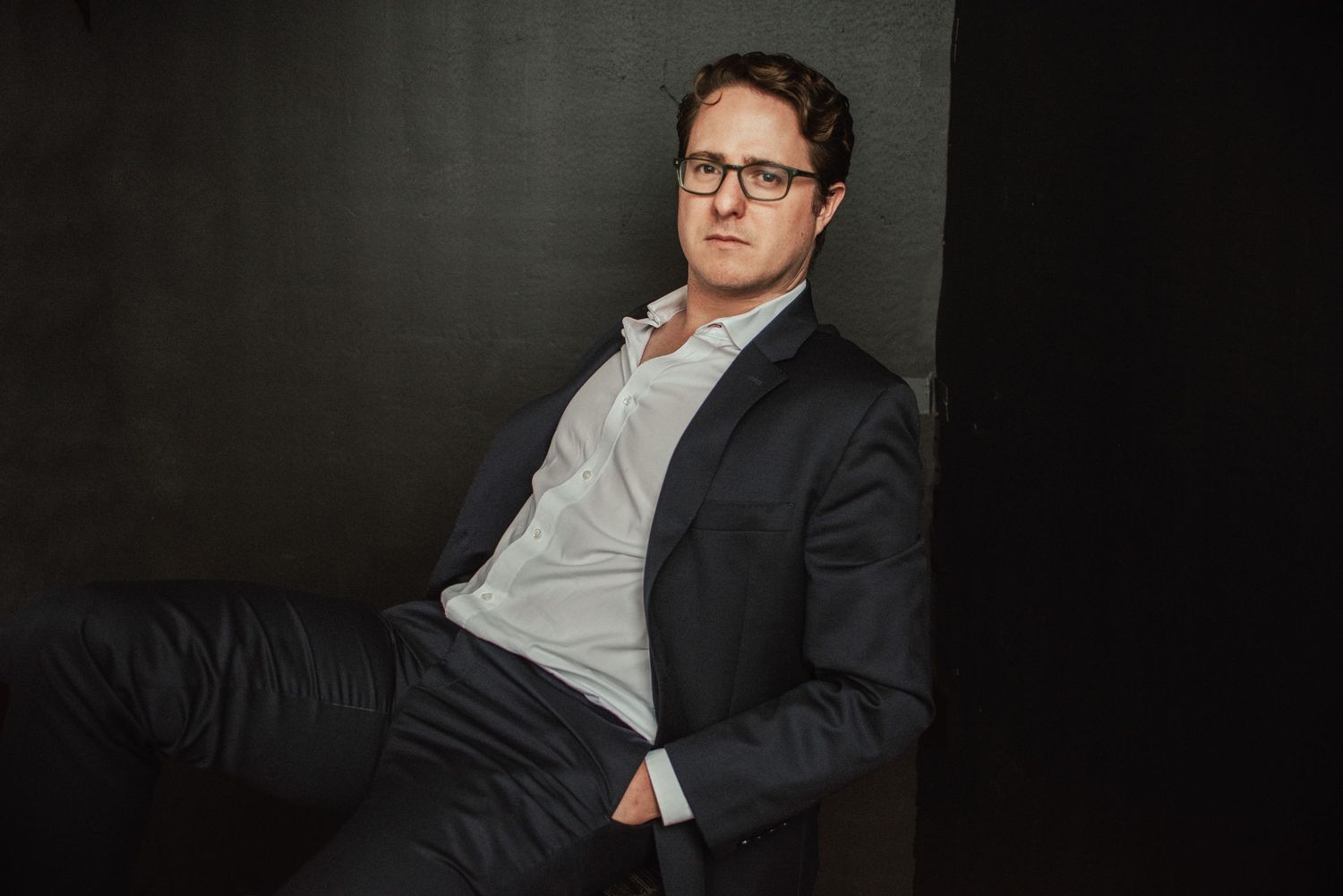 A person wearing glasses and a dark blazer poses against a black background in a professional photoshoot.