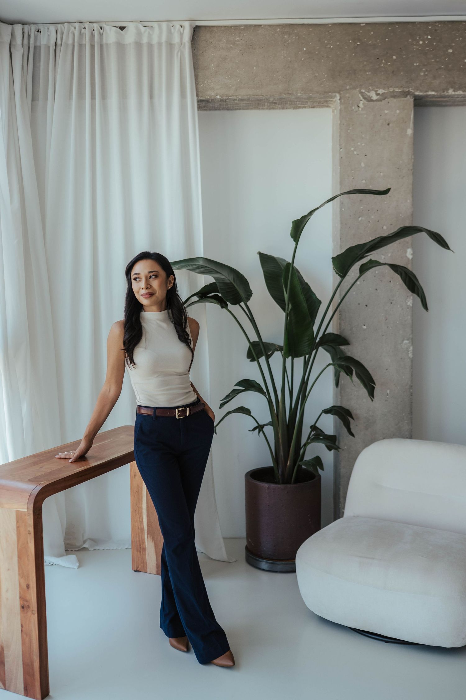 Woman in beige top and navy flared pants leaning on wooden table beside tropical plant in modern white interior.