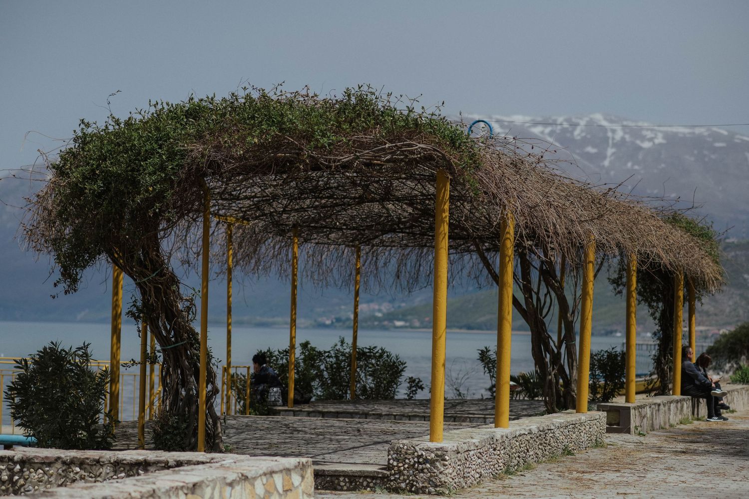 A panoramic view of covered pergolas with yellow posts along a stone wall against snow-capped mountains in the distance.