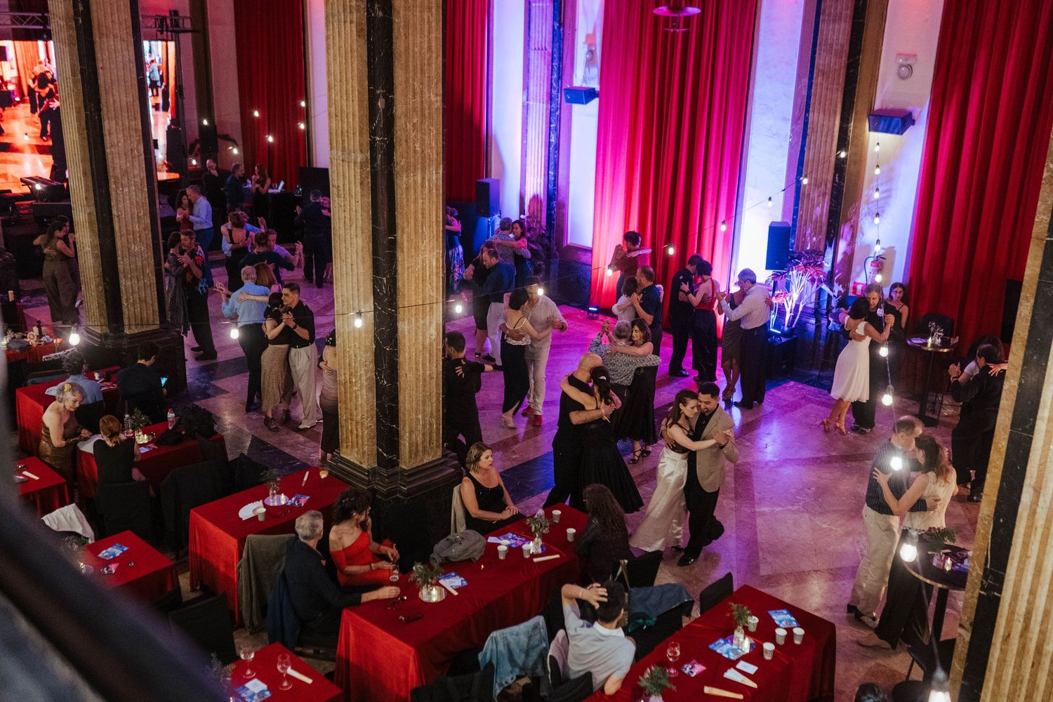 Elegant ballroom event with couples dancing, red-draped tables, tall columns, and dramatic red lighting in a grand venue.