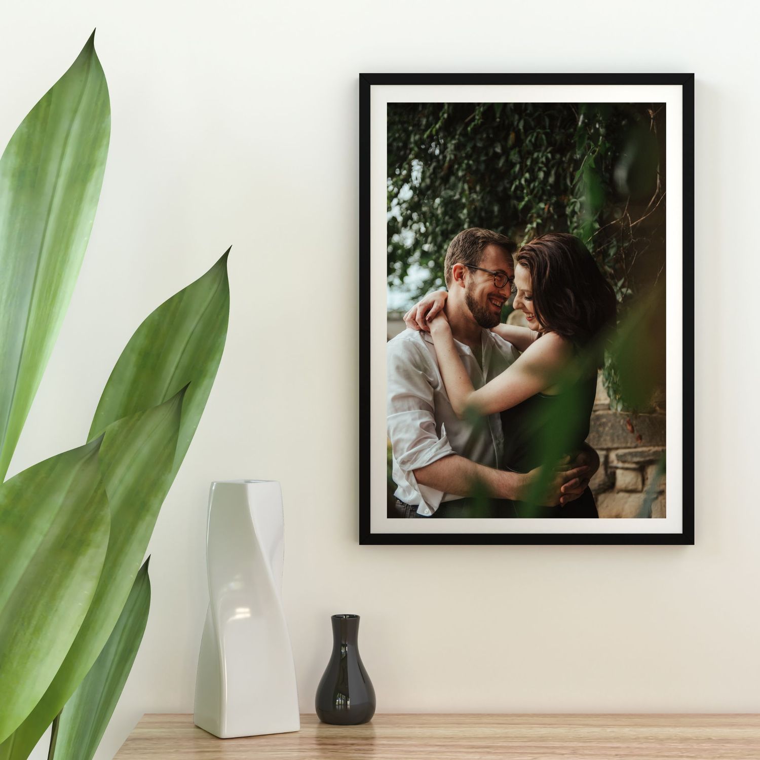 Indoor plant leaves frame a series of framed photographs displayed on a white wall above wooden furniture.