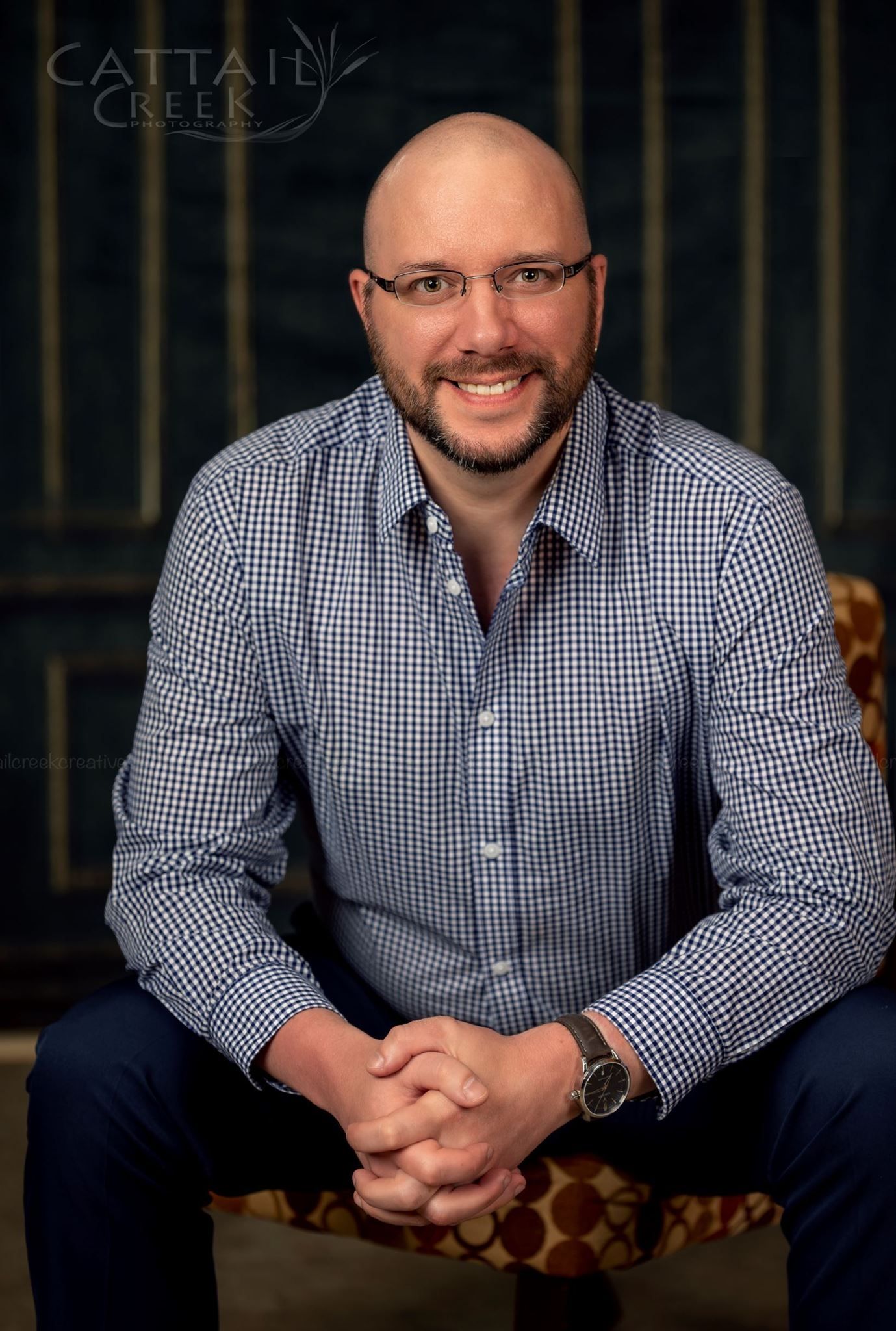 Professional headshot of someone in a checkered button-down shirt and glasses sitting on a patterned chair.