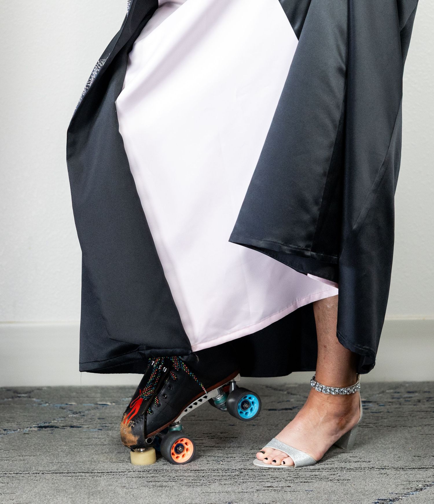 Person wearing a long black and white gown with one foot in a roller skate and the other in a clear heeled sandal.