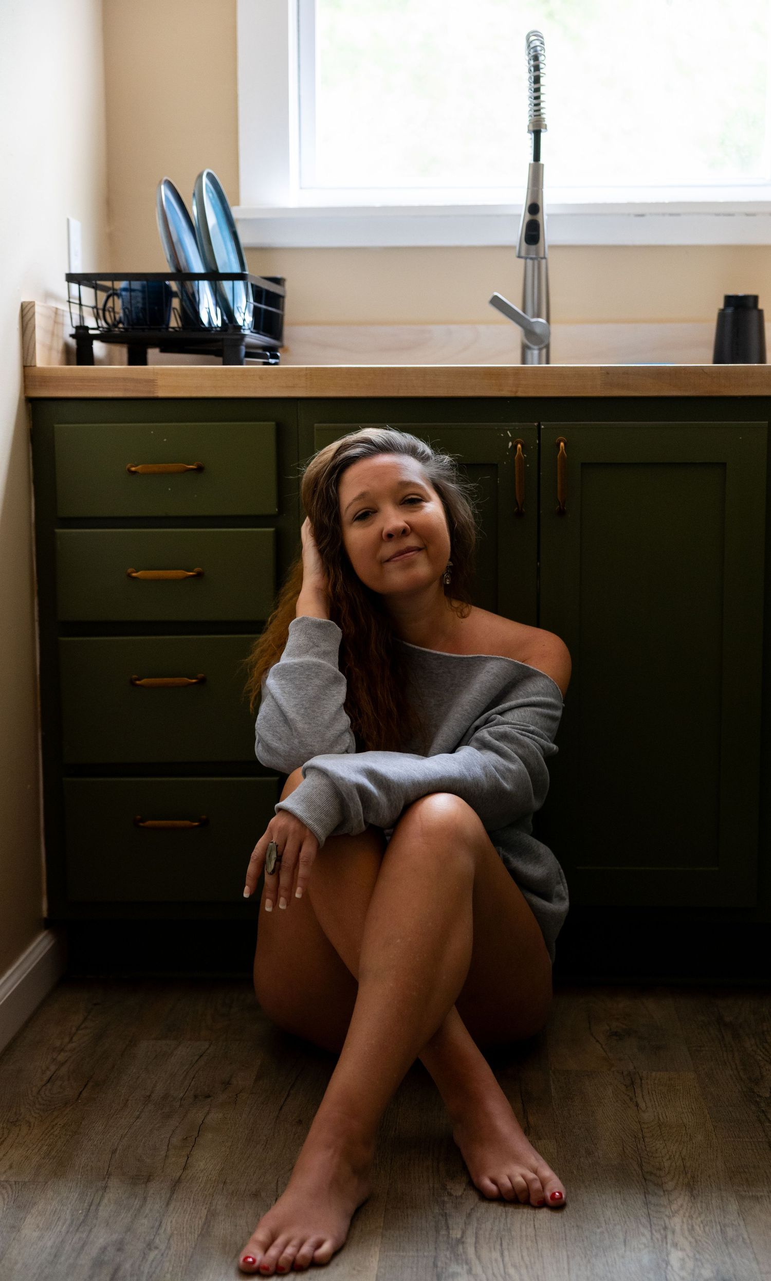 Woman sitting barefoot on kitchen floor in gray off-shoulder sweater beside green cabinets and wooden countertop.