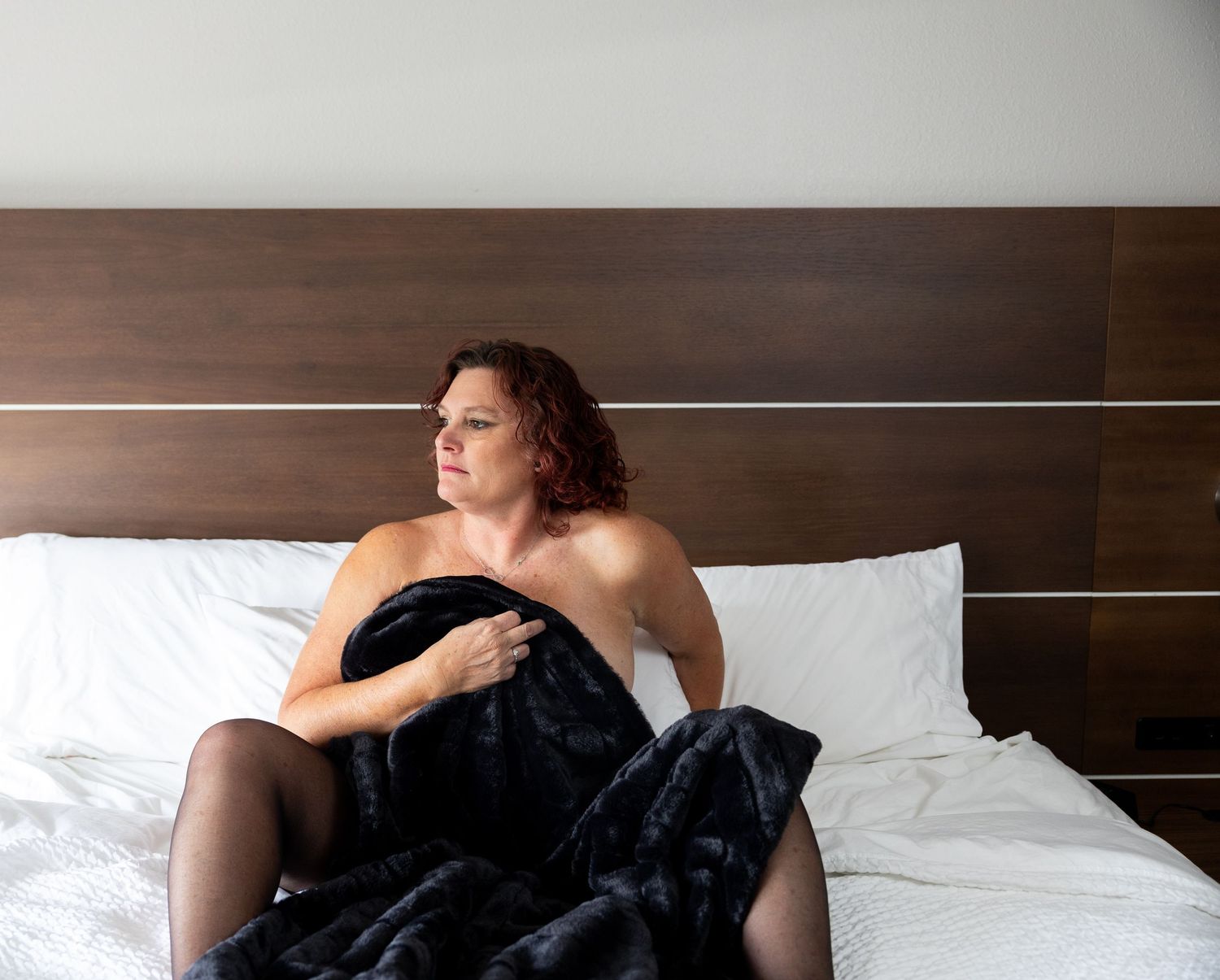 Woman wrapped in black fur blanket sitting on white bed with wooden headboard in hotel room.