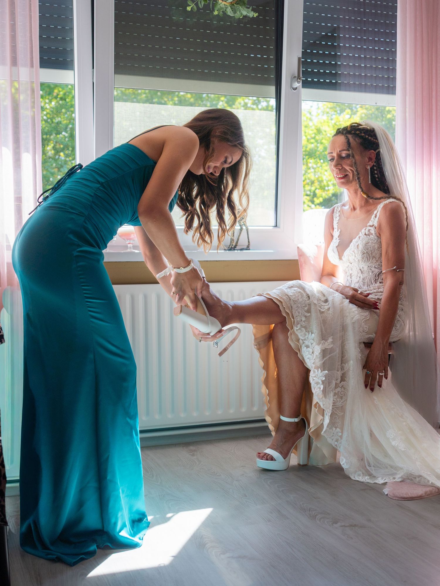 Woman in elegant teal gown helps adjust shoe on bride wearing lace wedding dress in sunlit room.