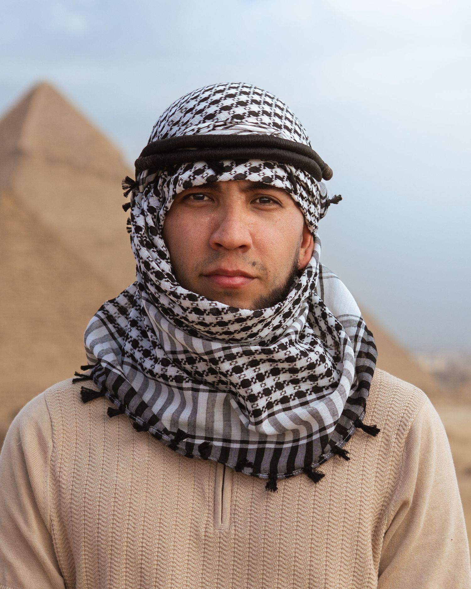 Someone wearing a traditional keffiyeh headscarf and sweater stands in front of the Egyptian pyramids in Giza.