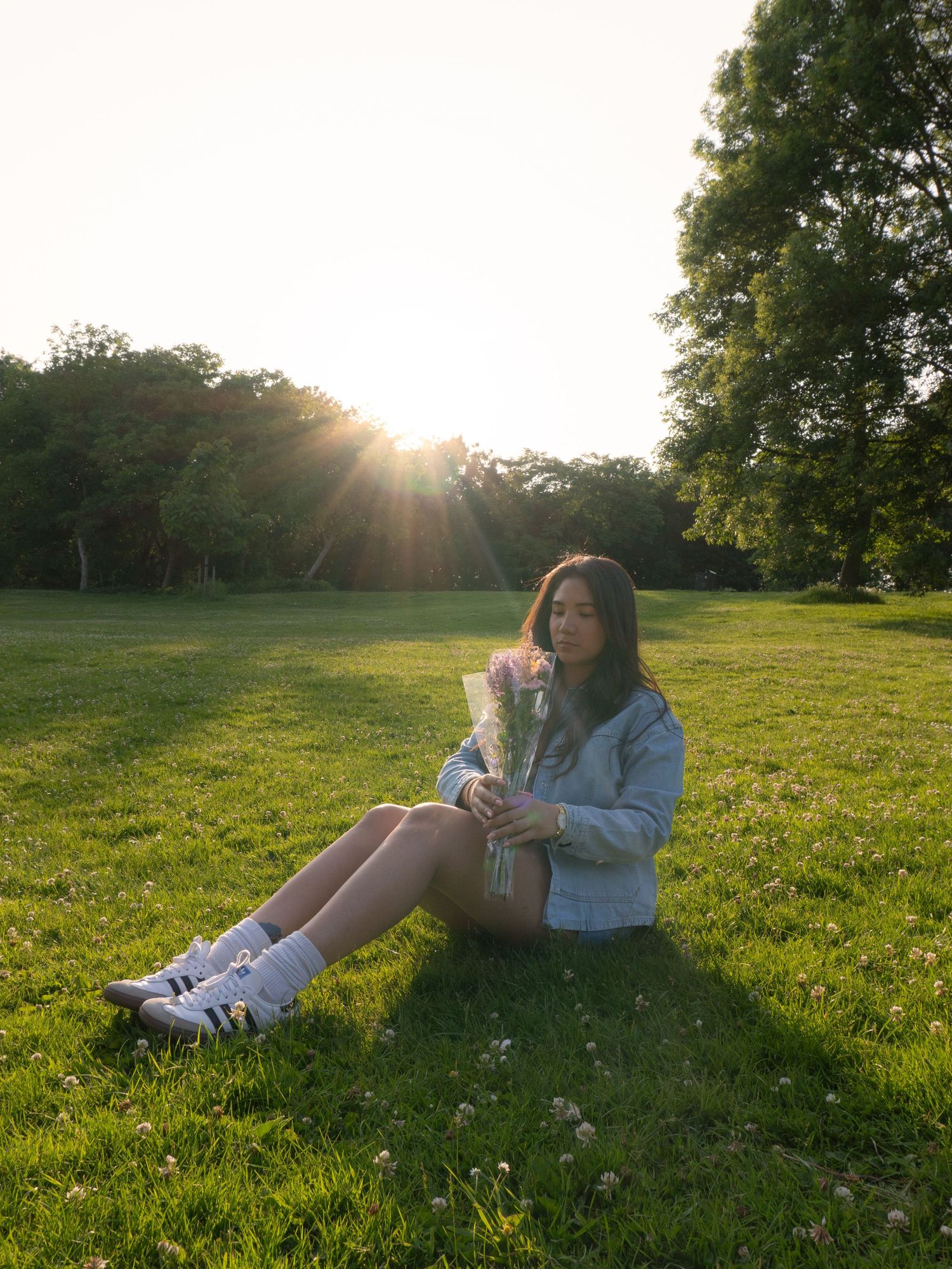 Someone sits on grass in a park at sunset wearing denim jacket and sneakers while enjoying the golden hour.