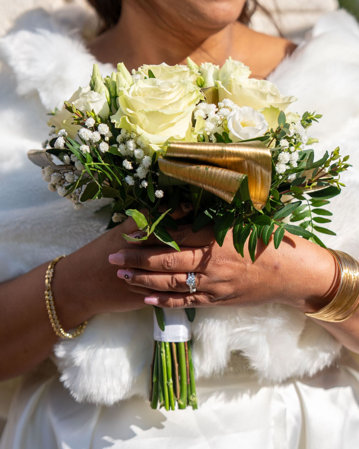 Close-up of bridal bouquet with white roses and greenery being held against a white wedding dress in natural light.