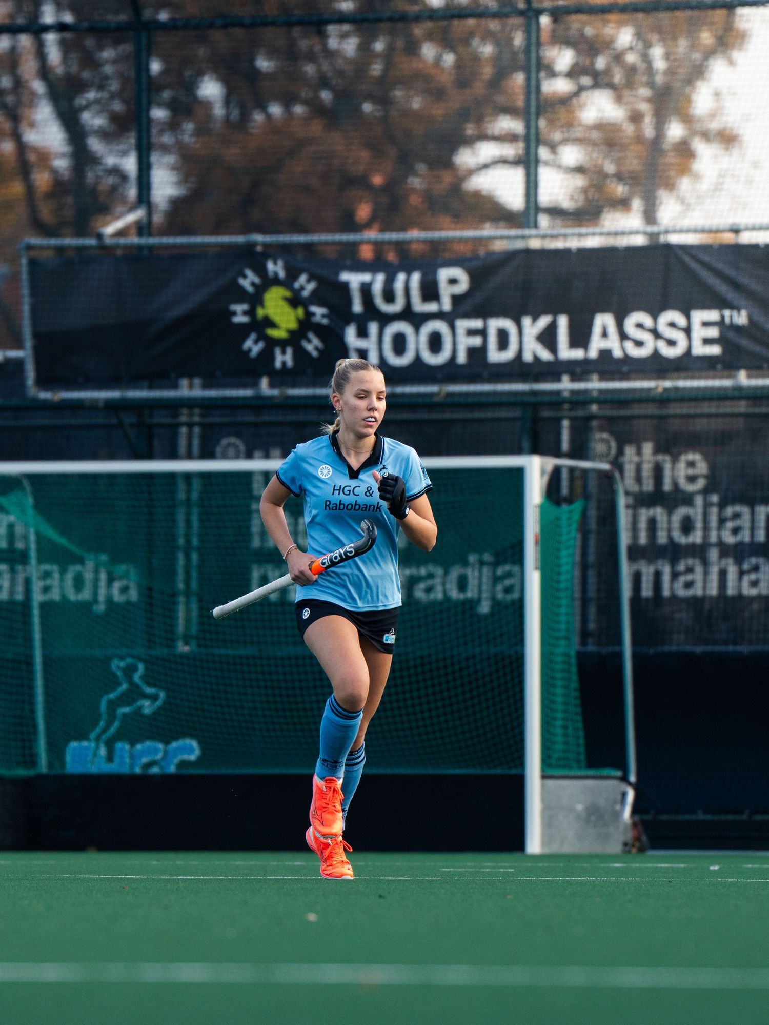 Field hockey player in light blue uniform performs a powerful shot during a match on artificial turf field.