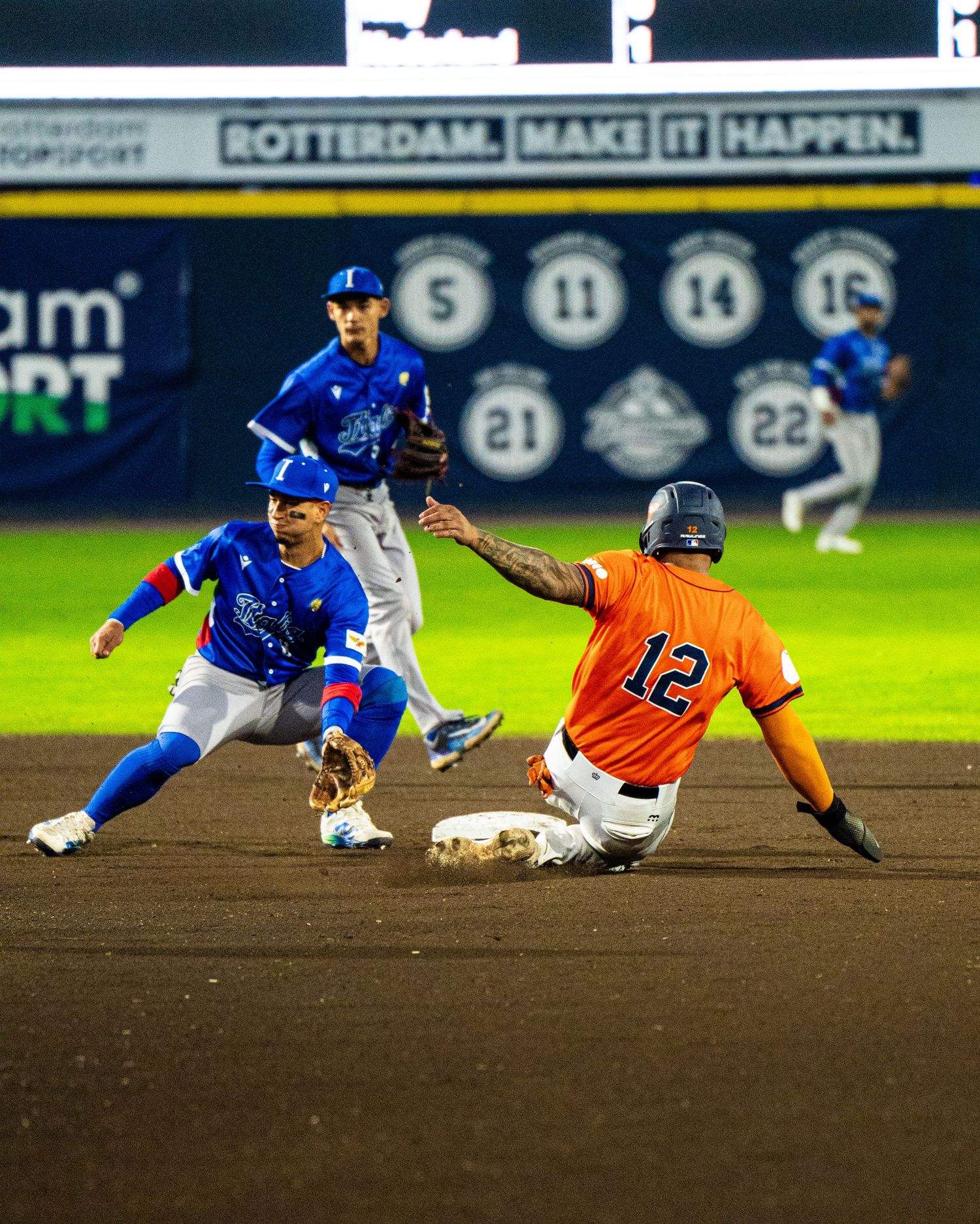 Baseball player in orange uniform slides into second base while blue team fielders attempt to make the play.