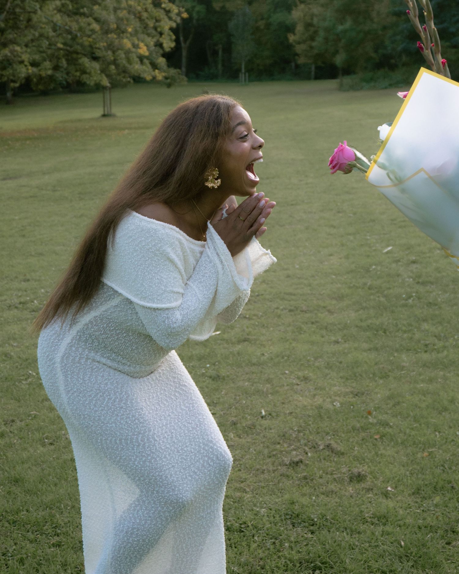 Someone in a white knit dress stands in a grassy outdoor setting with an animated expression.