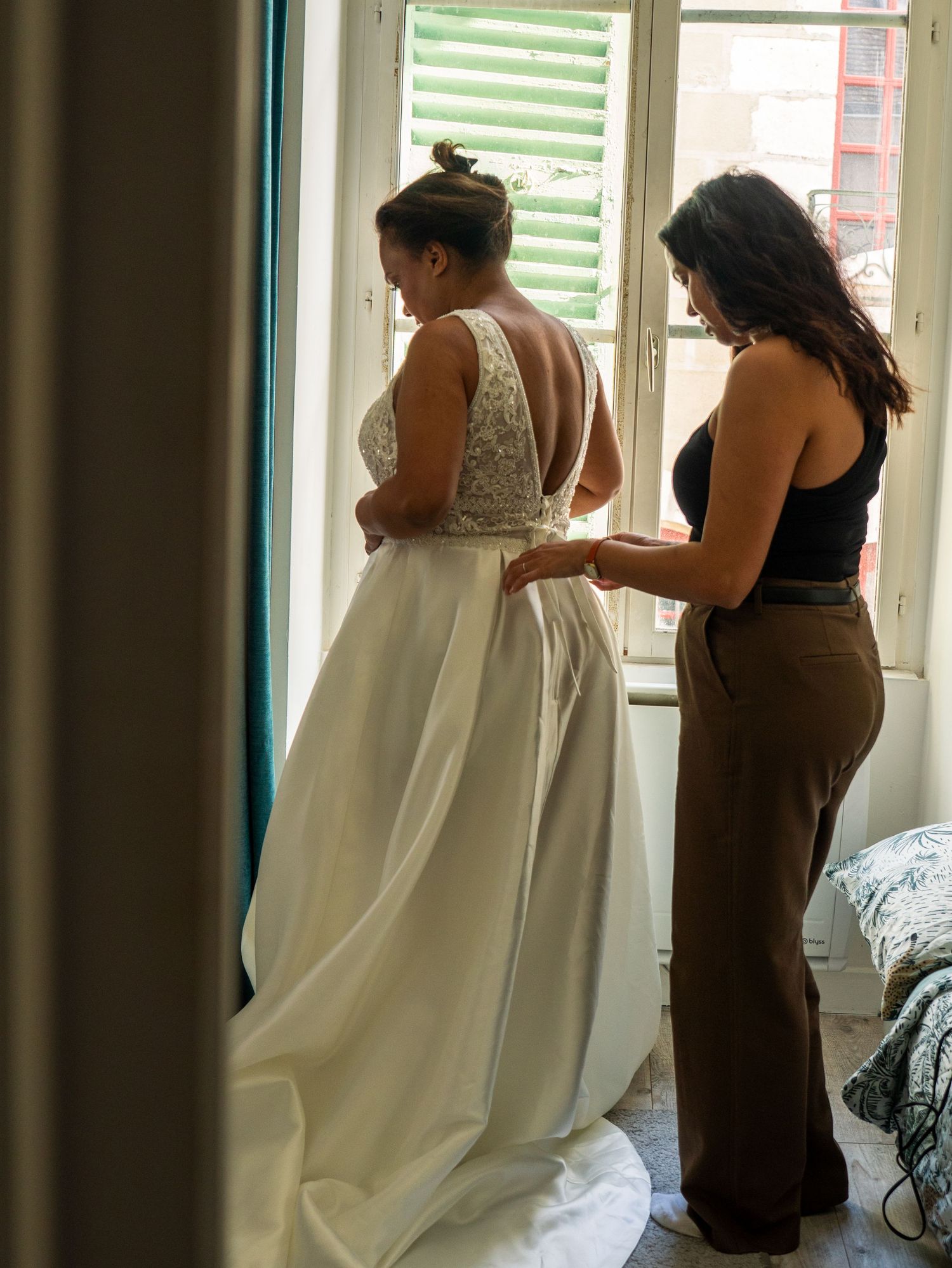 A series of wedding preparation photos showing someone being helped into a white wedding dress in a sunlit room.